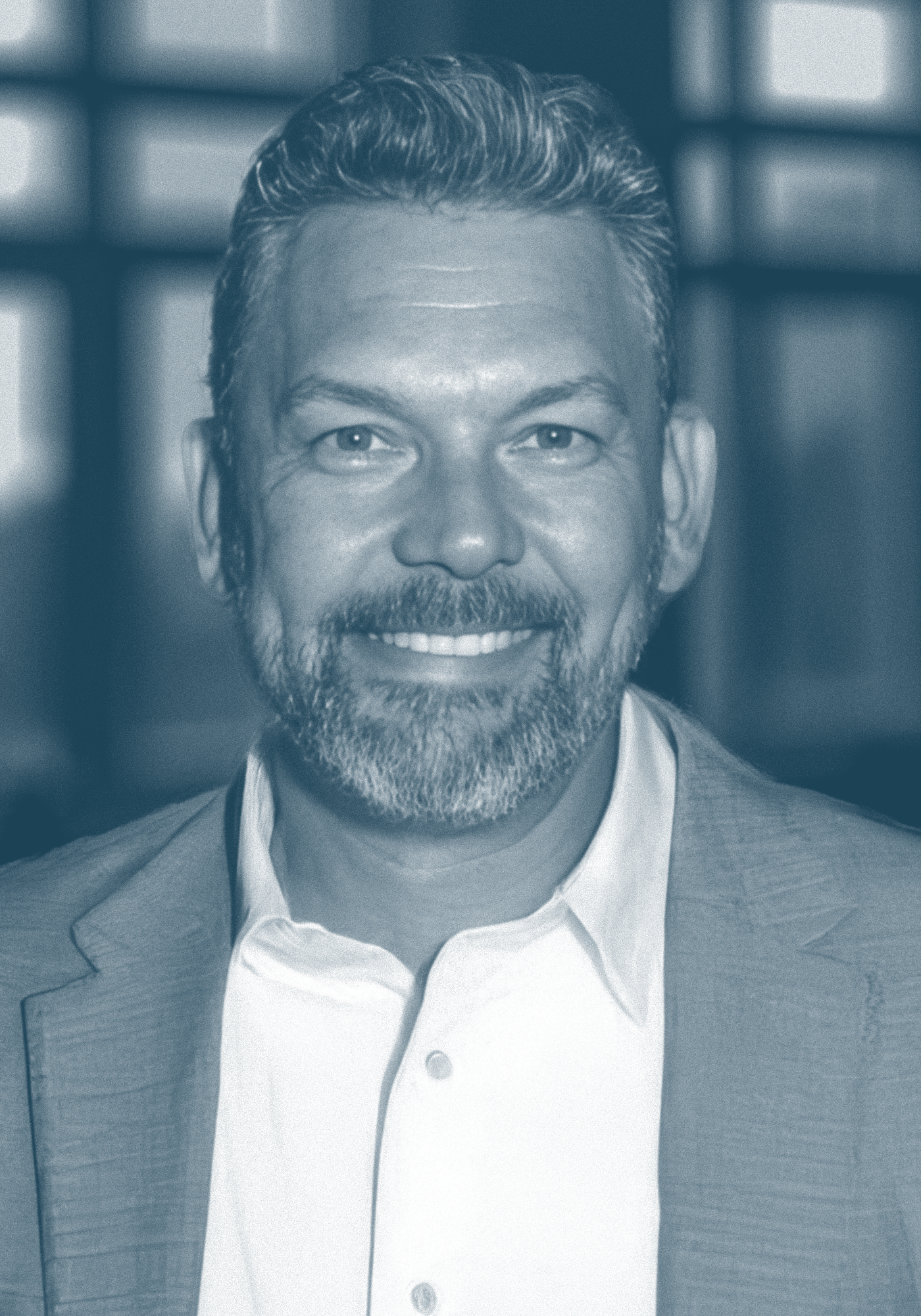 Close-up of a smiling man with gray hair and a beard, wearing a suit jacket and a white shirt, in front of a blurred background.