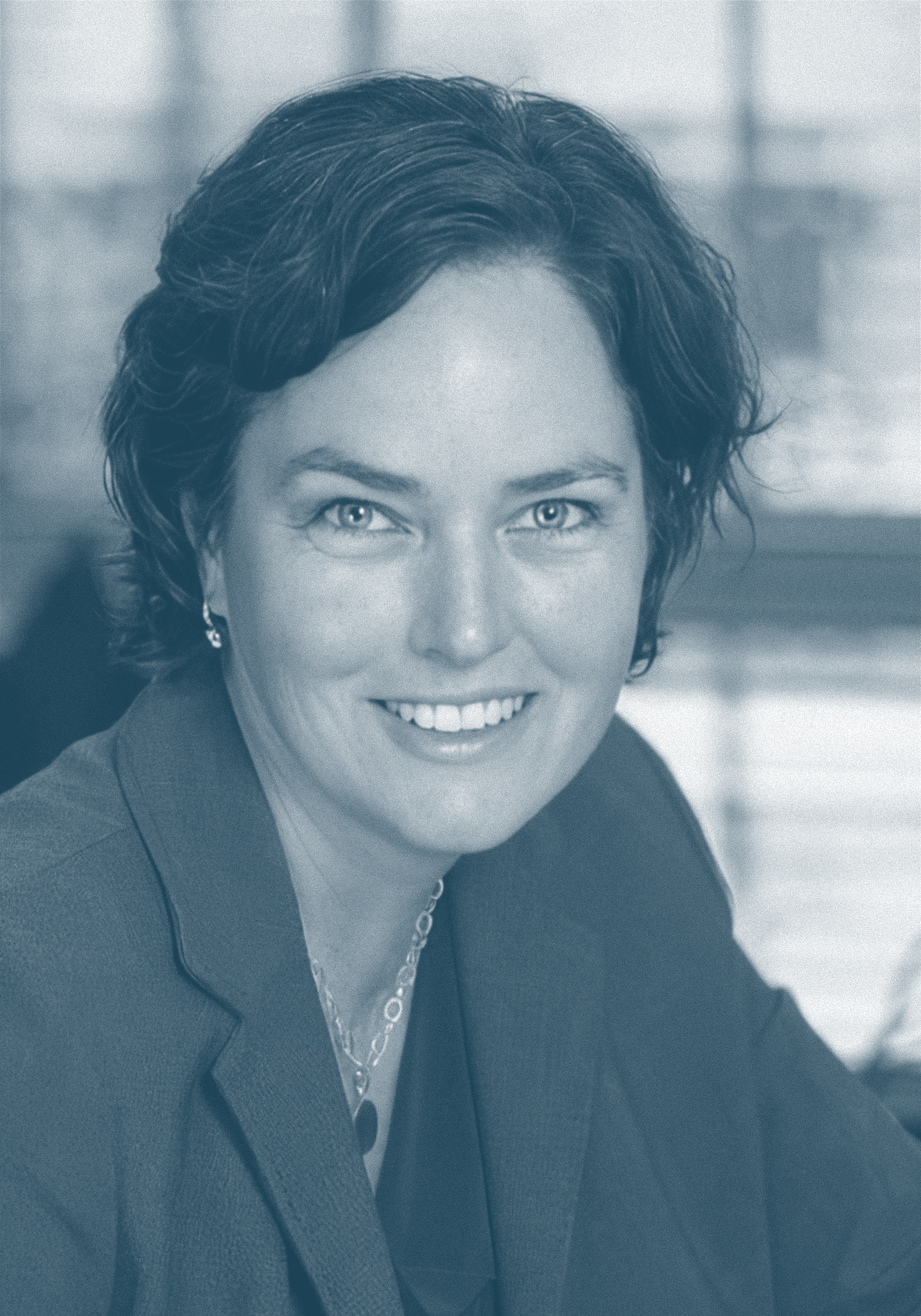 A woman with short curly hair, wearing a blazer and a necklace, smiling in an indoor setting.