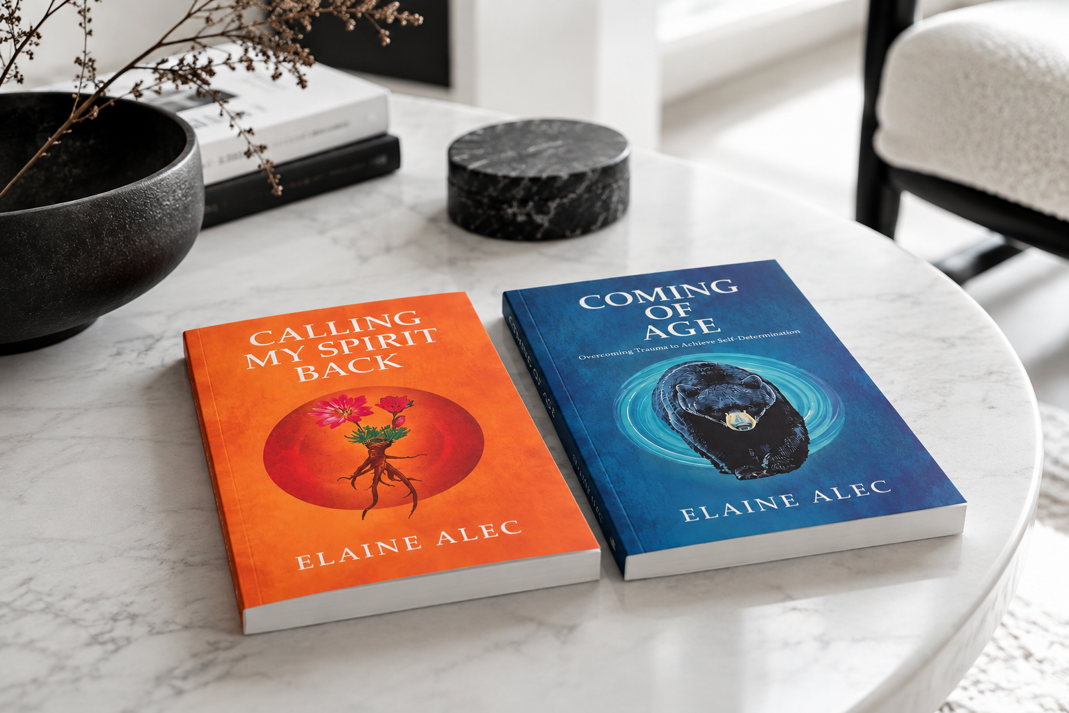 Two books titled "Calling My Spirit Back" and "Coming of Age" on a white marble table with a black bowl, black and white books, a black circular container, and a chair with a white cushion in the background.