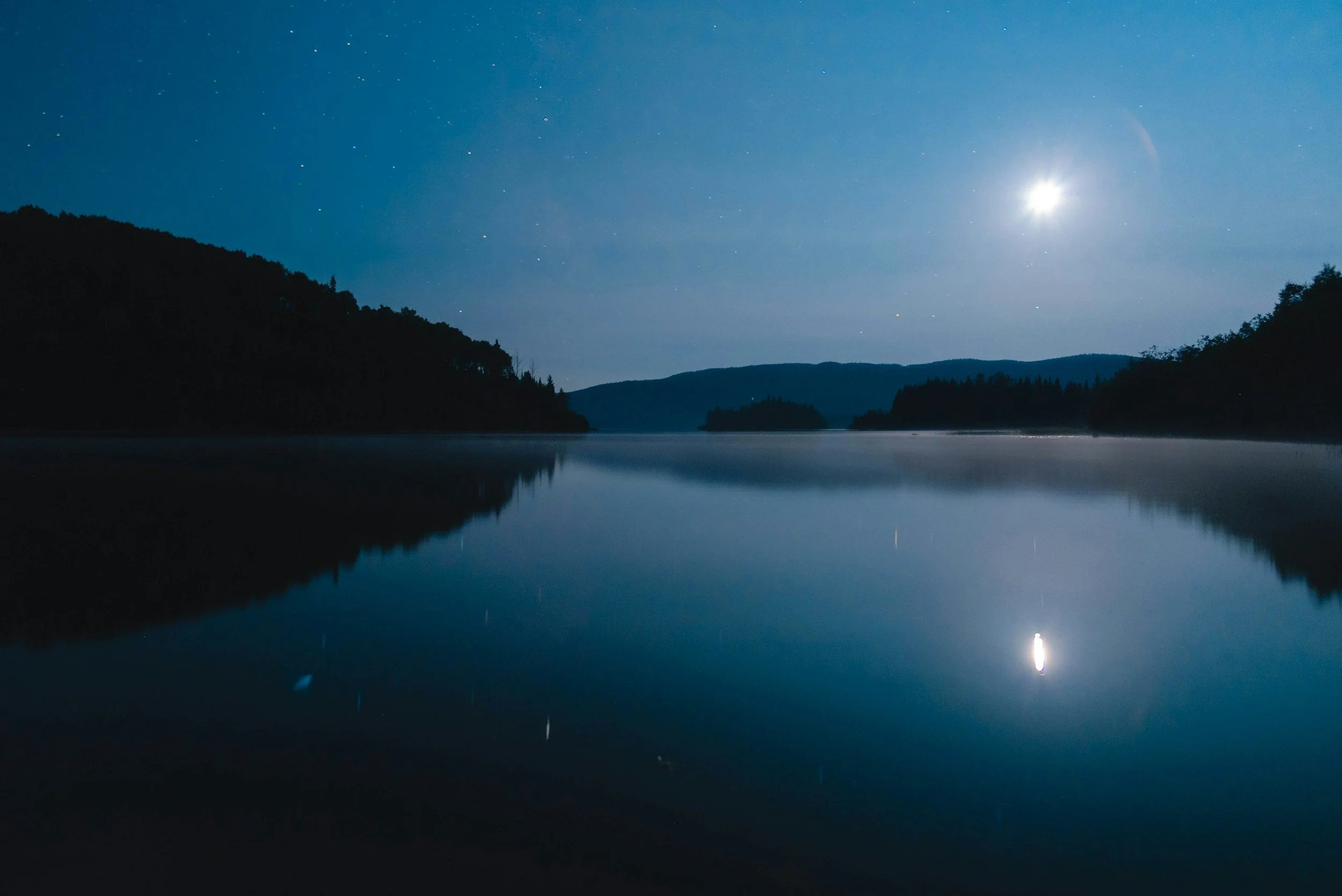Nighttime scene showing a calm lake with reflections of the moon and stars, silhouetted hills in the background.