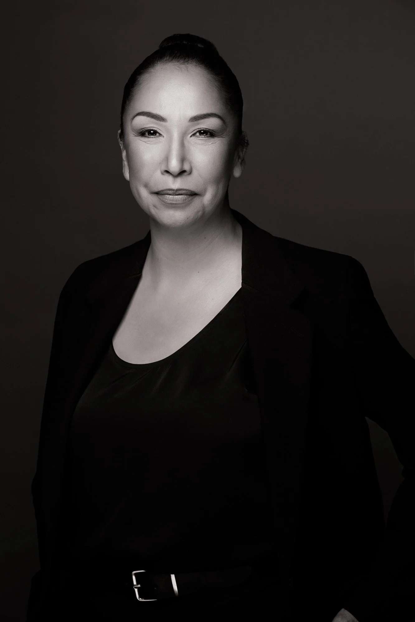 Black and white portrait of a smiling woman with hair styled in a bun, wearing a black blazer and shirt, against a dark background.