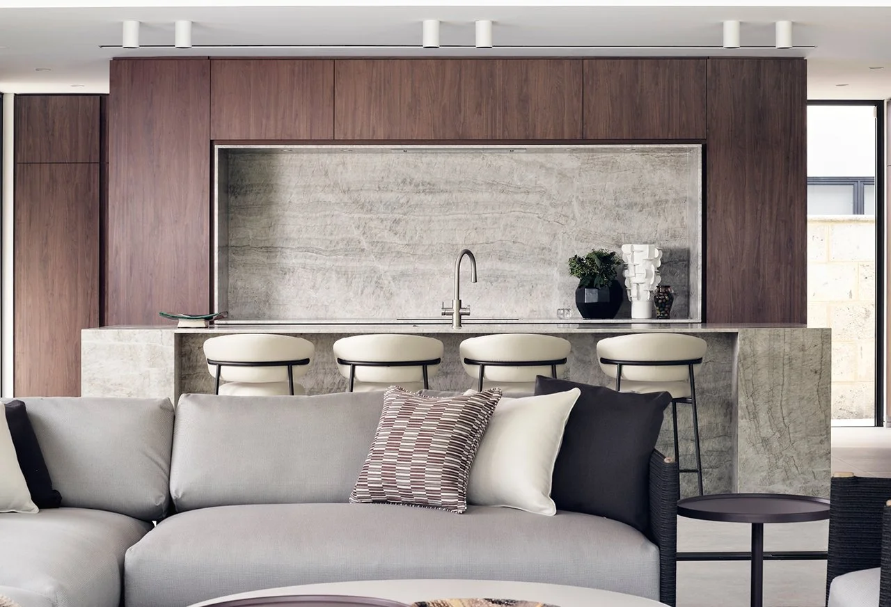 Modern living room with a beige sofa, decorative pillows, and a kitchen area with a marble island, brown wood cabinets, and barstools.