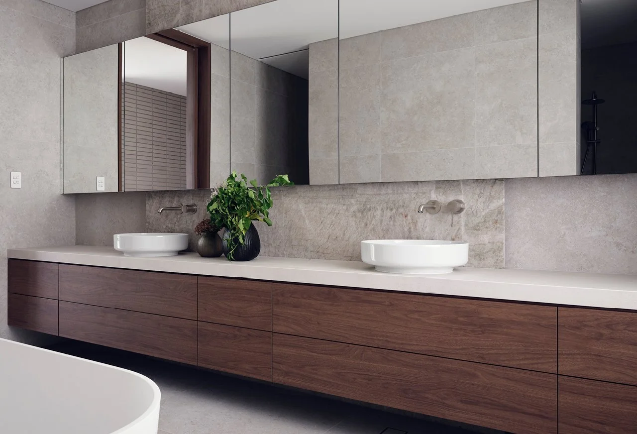 Modern bathroom vanity with double white vessel sinks, a large mirror, a wooden cabinet, and a vase with green plants.