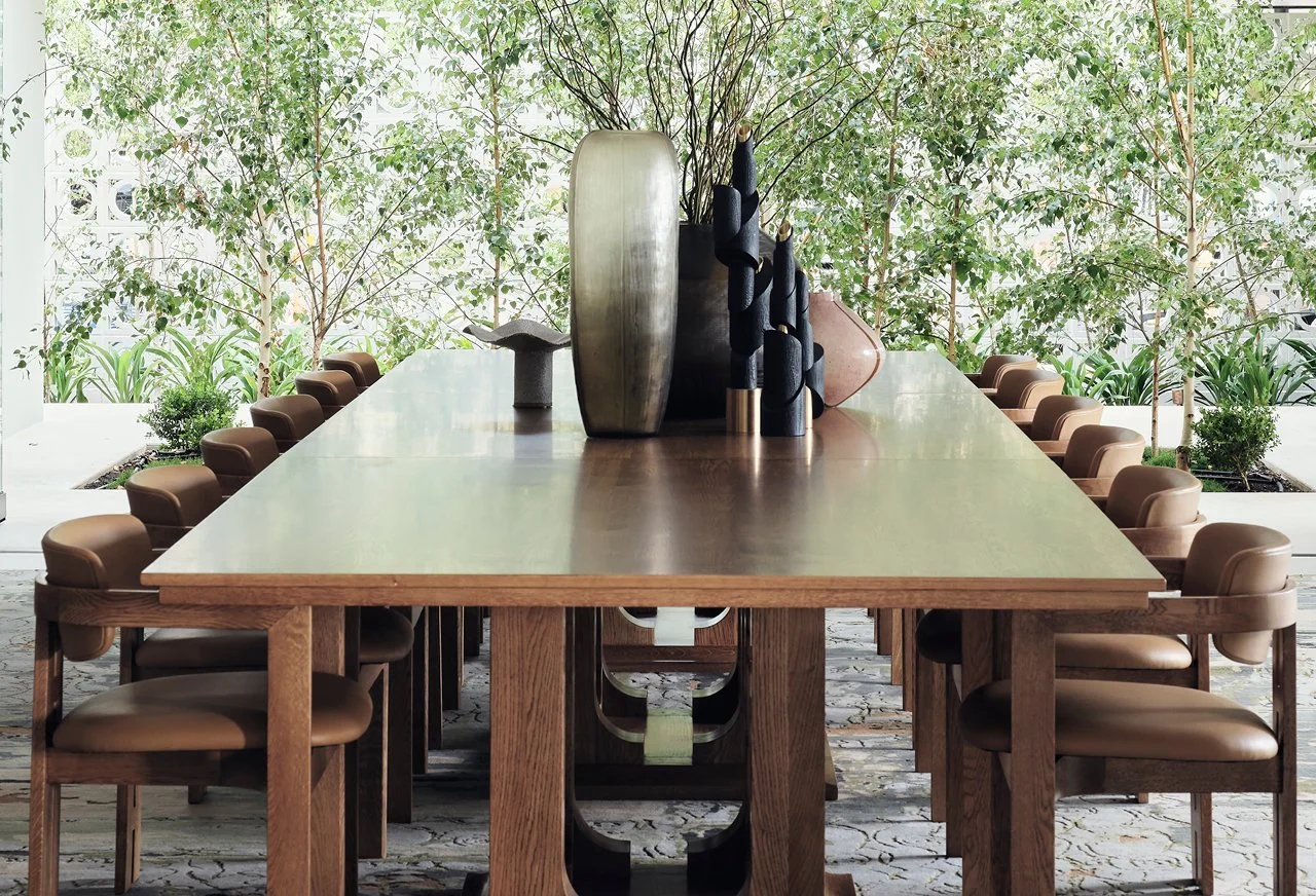 A long wooden dining table with beige cushioned chairs on both sides, set in a bright room with large windows and greenery outside. The table has decorative vases and sculptures.