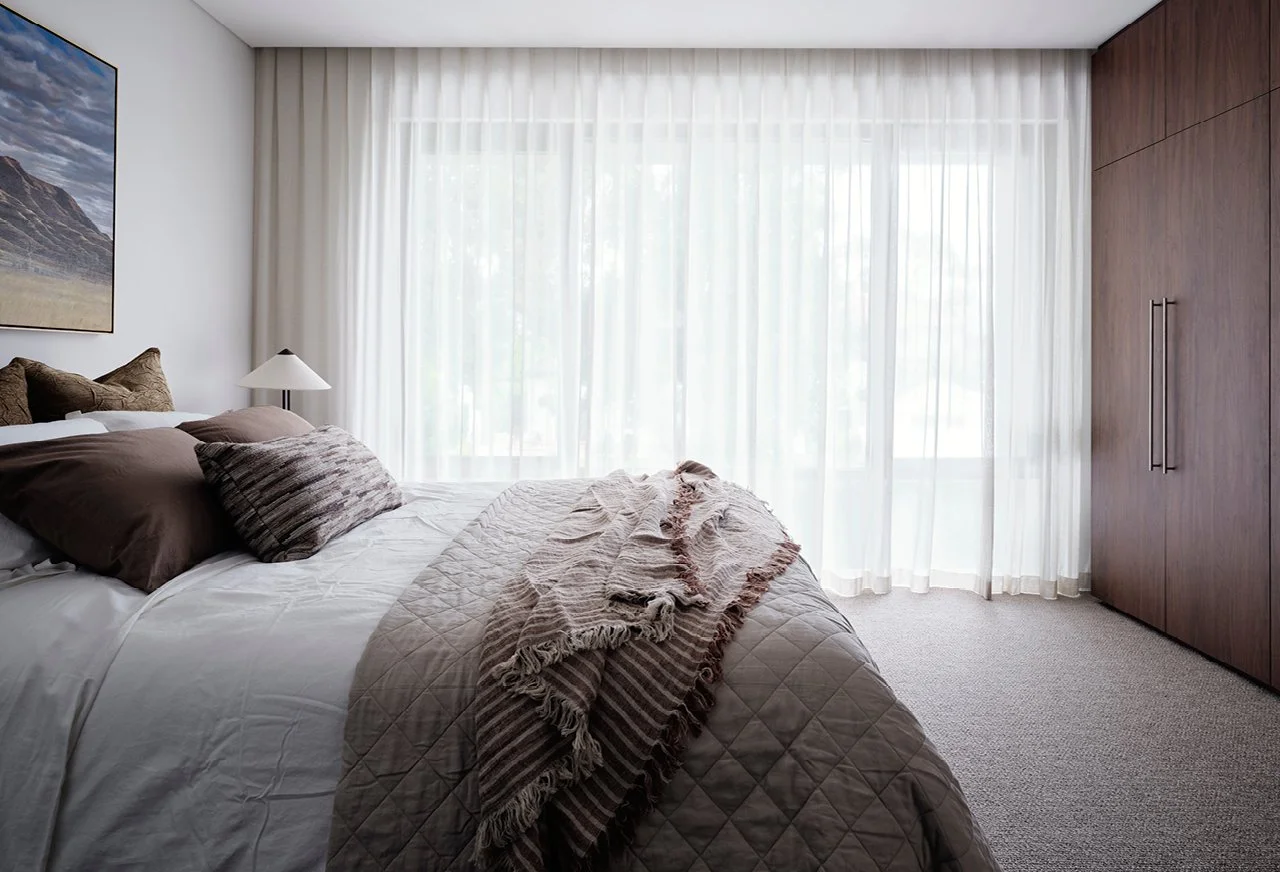 A neatly made bedroom with a bed, pillows, quilt, and a throw blanket, large window with sheer curtains, a nightstand with a lamp, and a wooden closet.