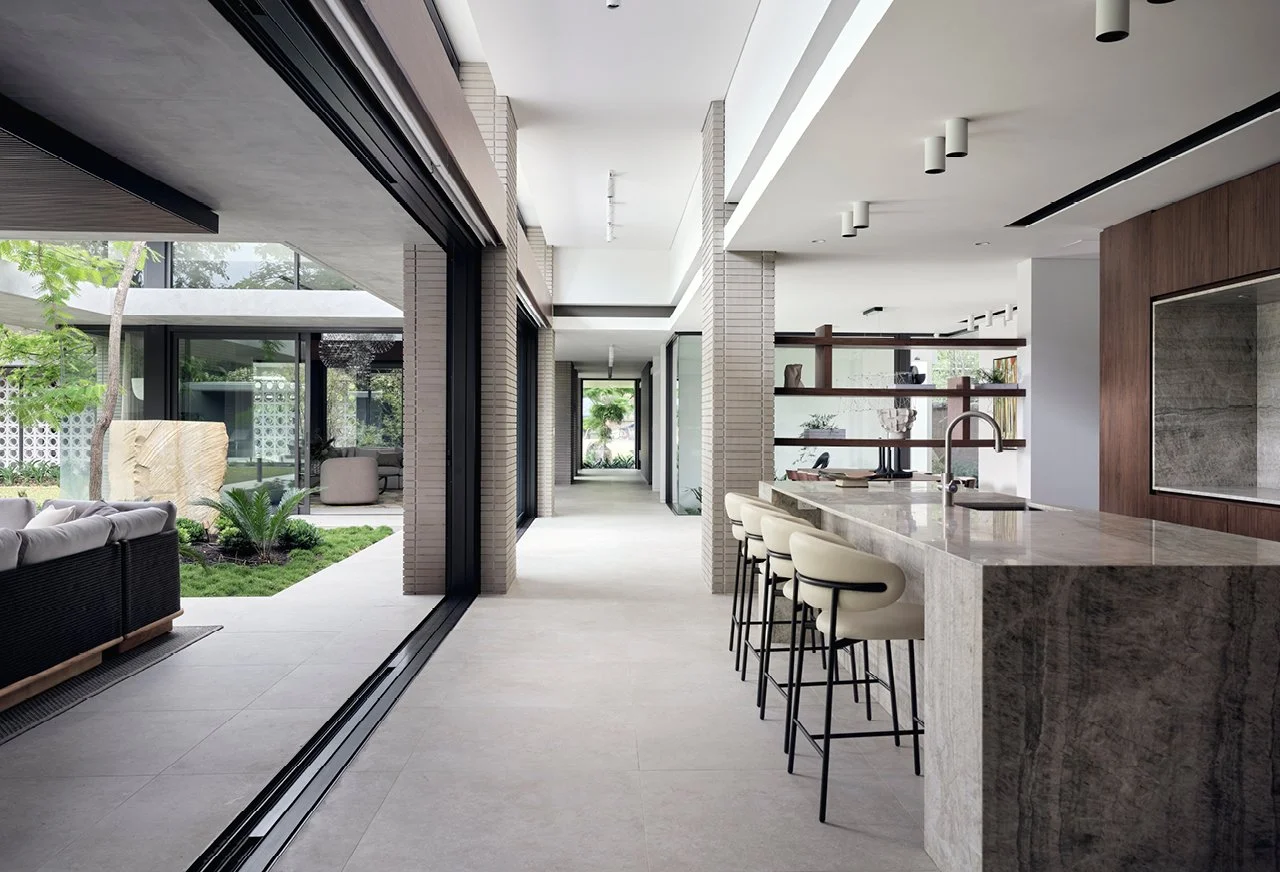 Modern open-concept kitchen with a marble island and barstools, adjacent to a living area with outdoor views and a garden.