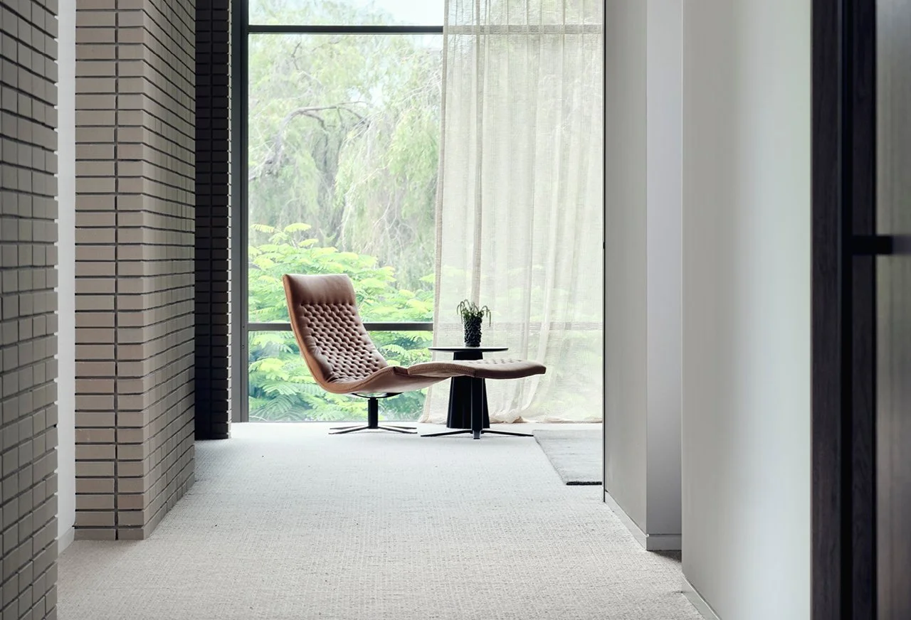 Modern interior with a tan lounge chair and small table with a potted plant near large floor-to-ceiling window with sheer curtains, overlooking greenery.