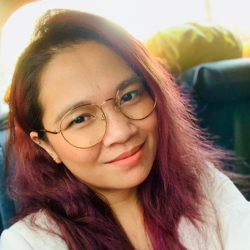 Close-up of a young woman with glasses, pink and purple hair, smiling.