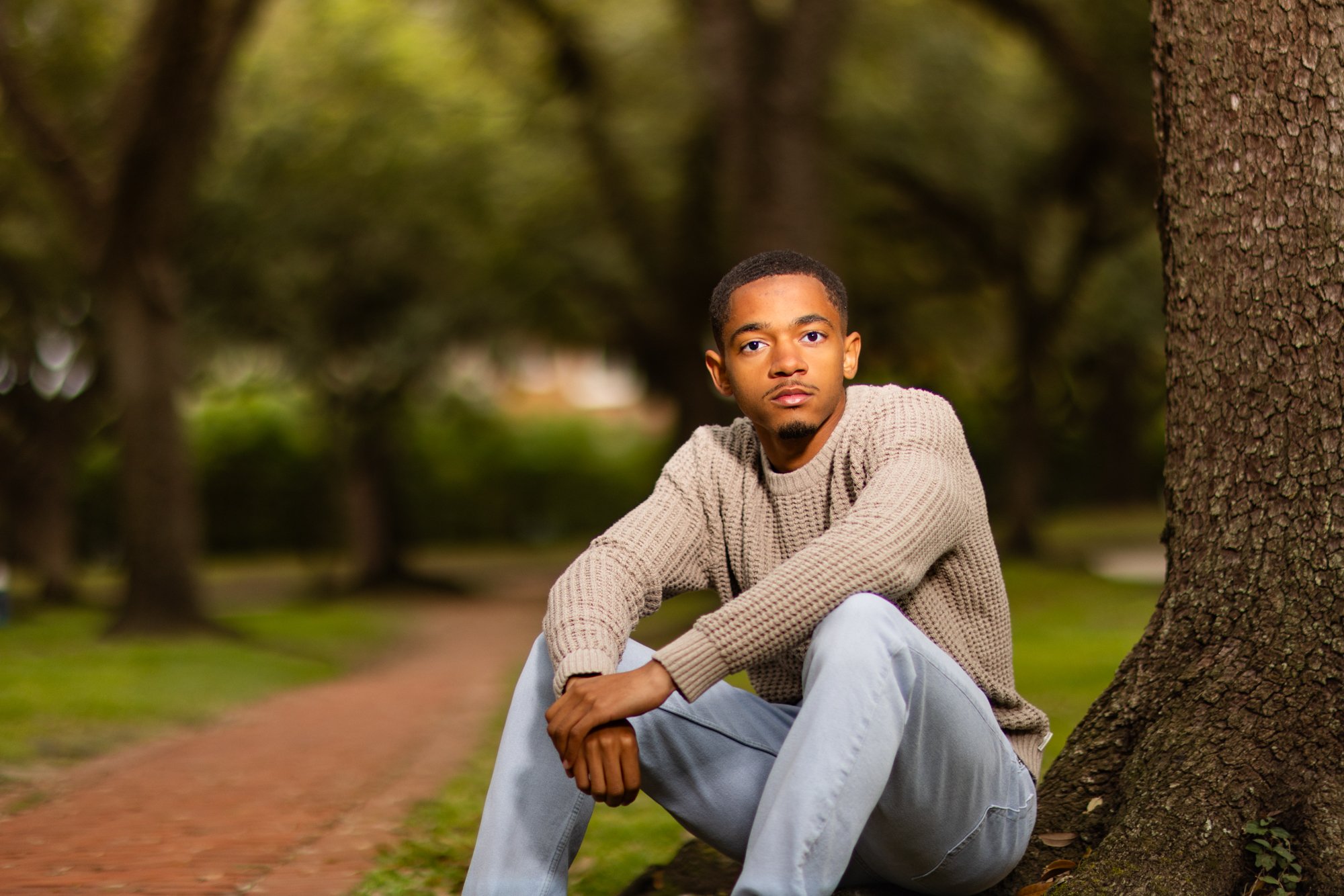 A young man with short dark hair, wearing a beige sweater and light gray pants, sitting outdoors next to a large tree in a park. He is looking directly at the camera with a neutral expression. The background shows blurred green trees and a pathway.