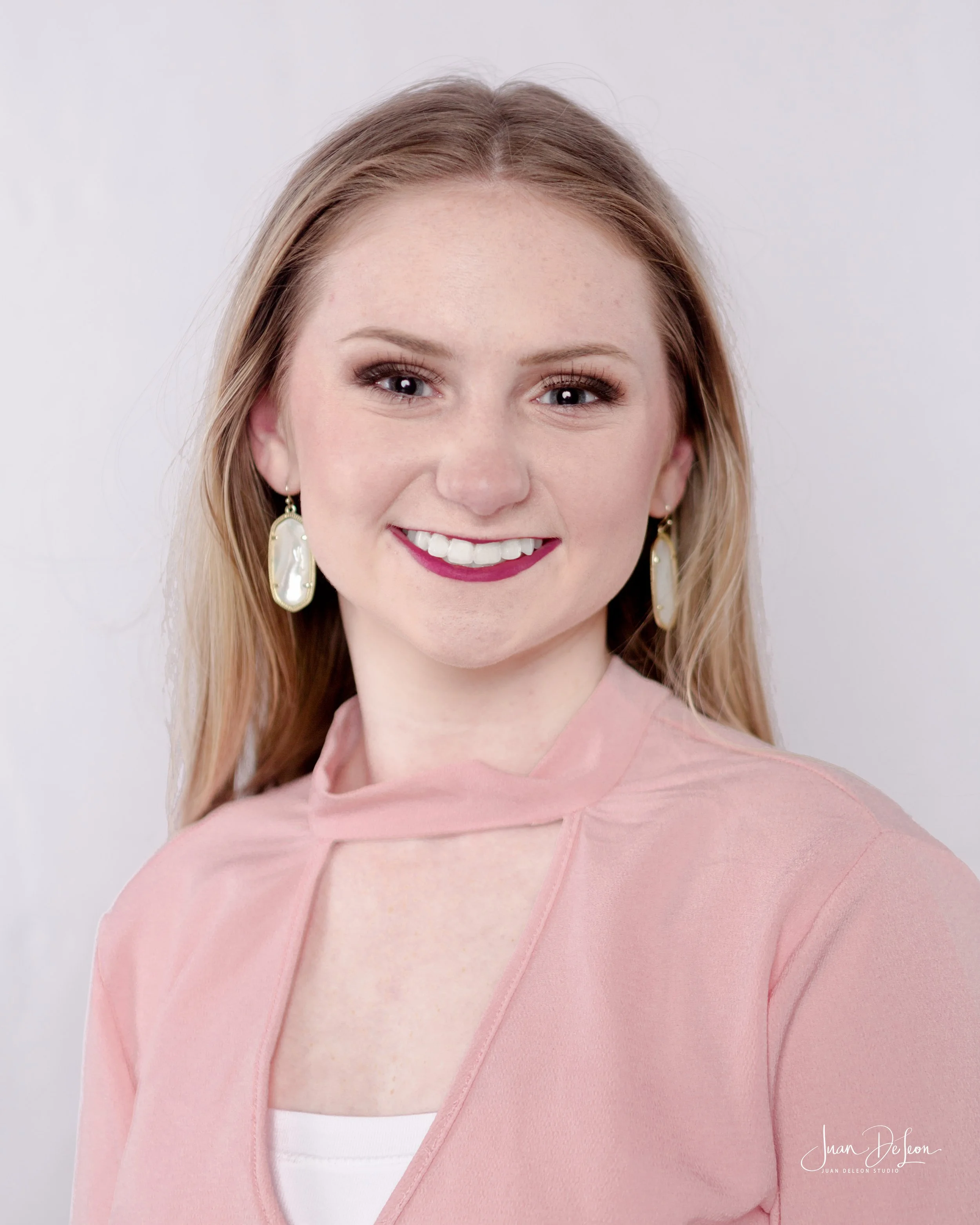 Close-up of a woman with blonde hair, smiling, wearing earrings and a light pink top with a white inner shirt, against a white background.