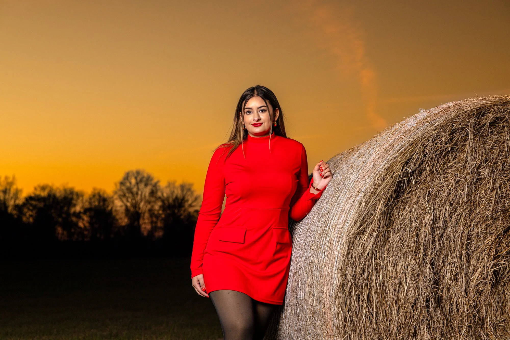 A woman in a red dress and black tights standing next to a large hay bale at sunset, with a sunset sky and silhouetted trees in the background.