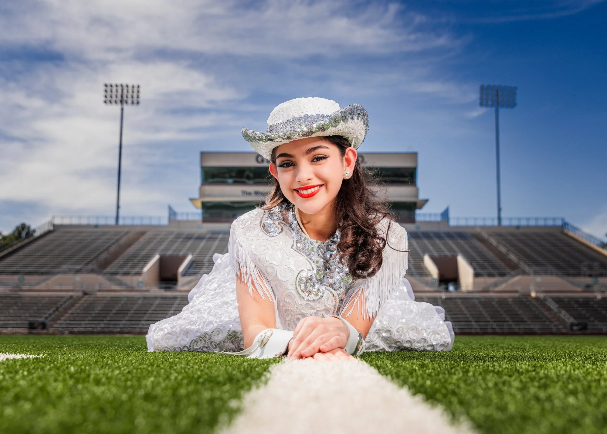 A young woman in a white dress and sequin cowboy hat lying on the football field at a stadium, smiling at the camera.