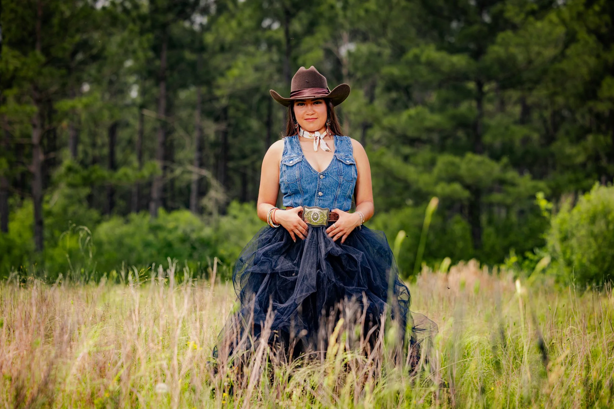 Woman in cowboy hat and denim vest standing in a field with trees in the background.