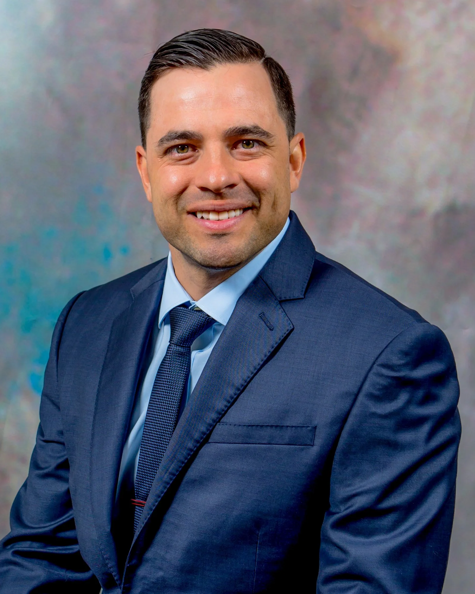 A man with short, dark hair and wearing a dark blue suit, white shirt, and dark tie, smiling in front of a neutral background.