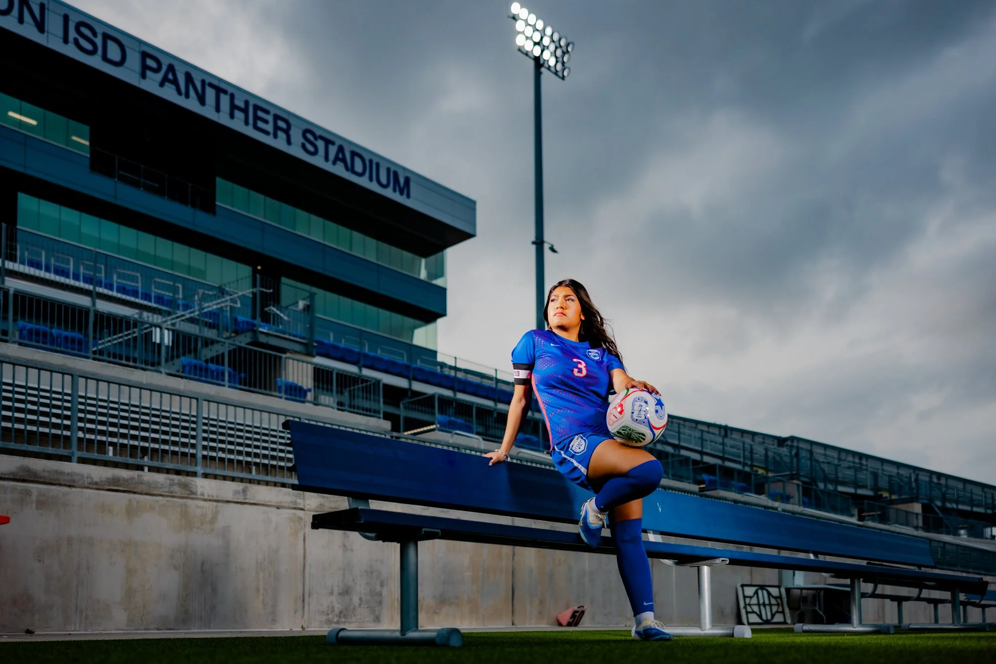 A female soccer player wearing a blue uniform and the number 3 sits on a bench in a stadium, holding a soccer ball, with a stadium building and floodlights in the background.