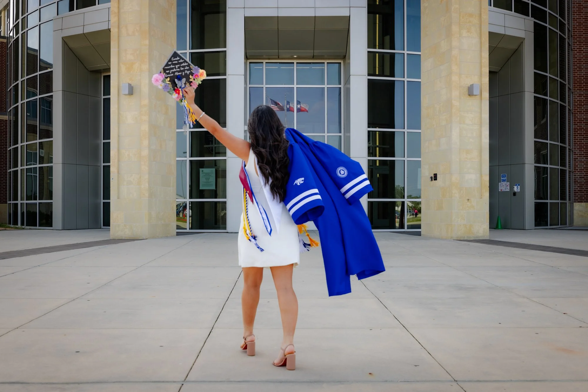 A woman in a white dress holding a decorated graduation cap in front of a modern building with large glass windows and flags, facing away from the camera.