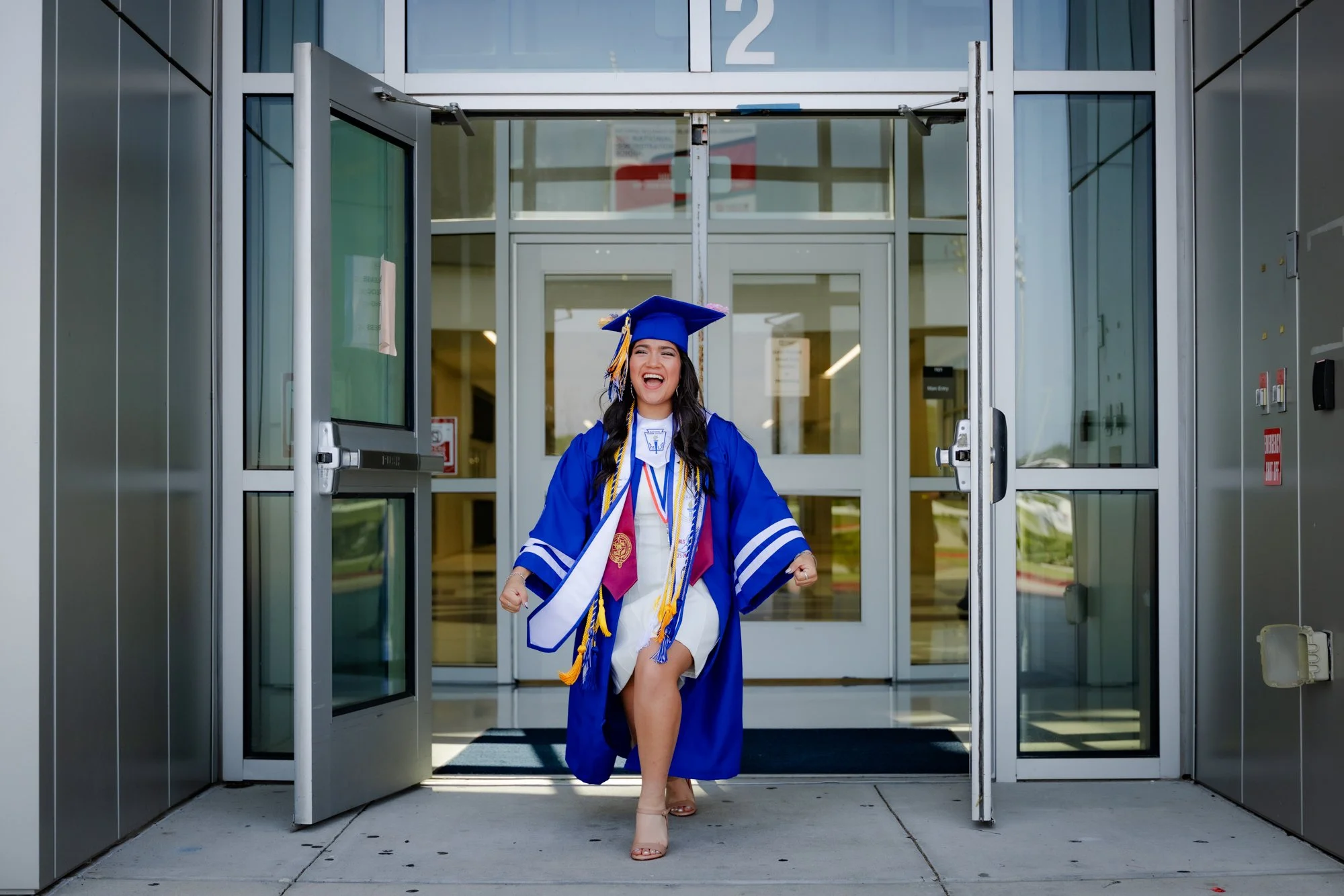 A young woman in a blue graduation cap and gown is smiling and walking out of a building through automatic glass doors, celebrating her graduation.