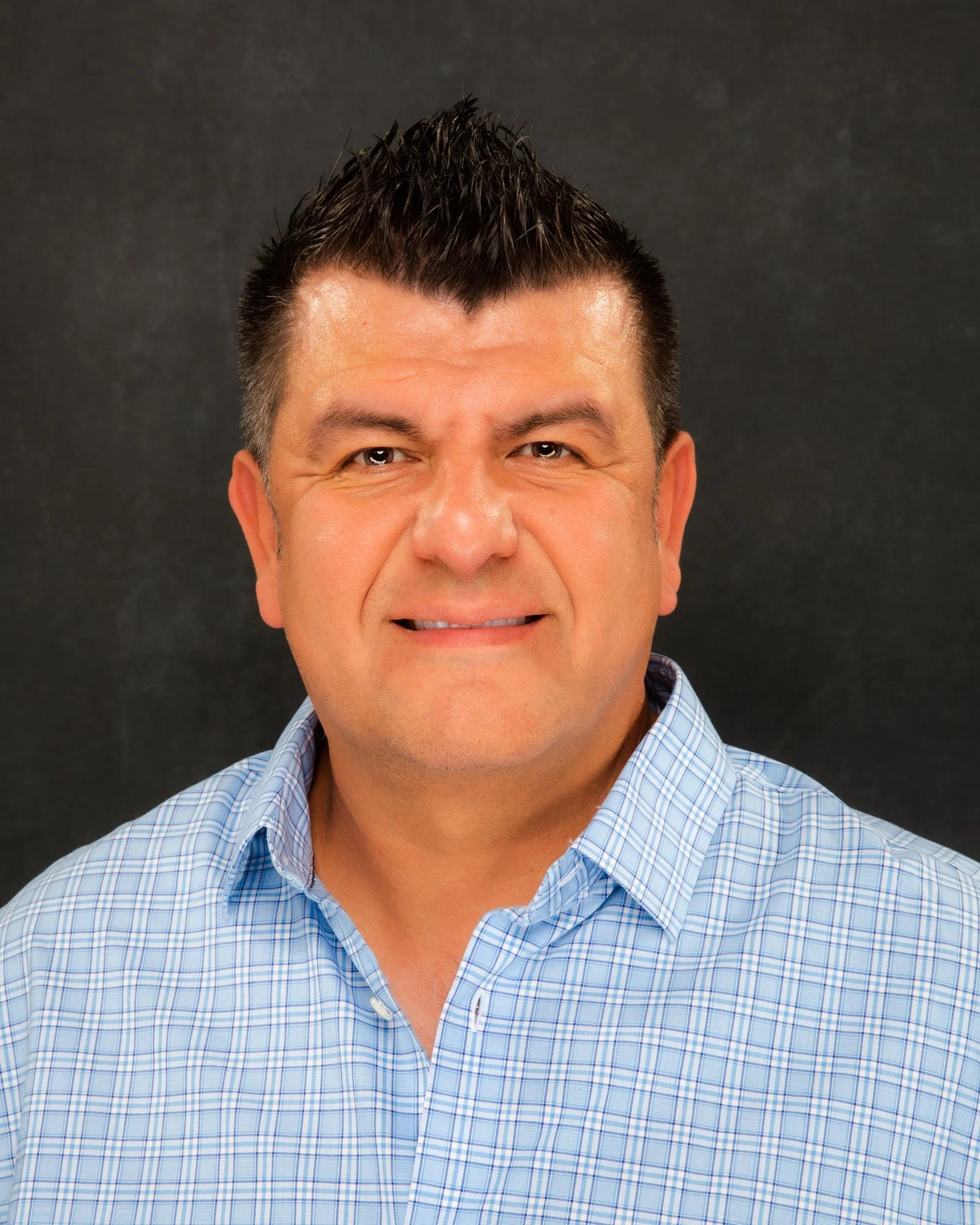 Close-up of a middle-aged man with dark, spiky hair and a slight smile, wearing a light blue checkered shirt, against a dark background.