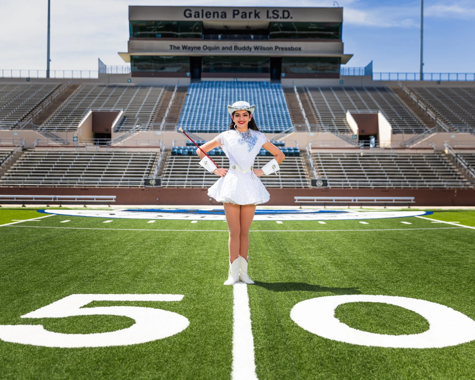 A young woman dressed in a white cowgirl costume holding a riding crop, standing in the middle of a football field with stadium seating and a press box in the background.