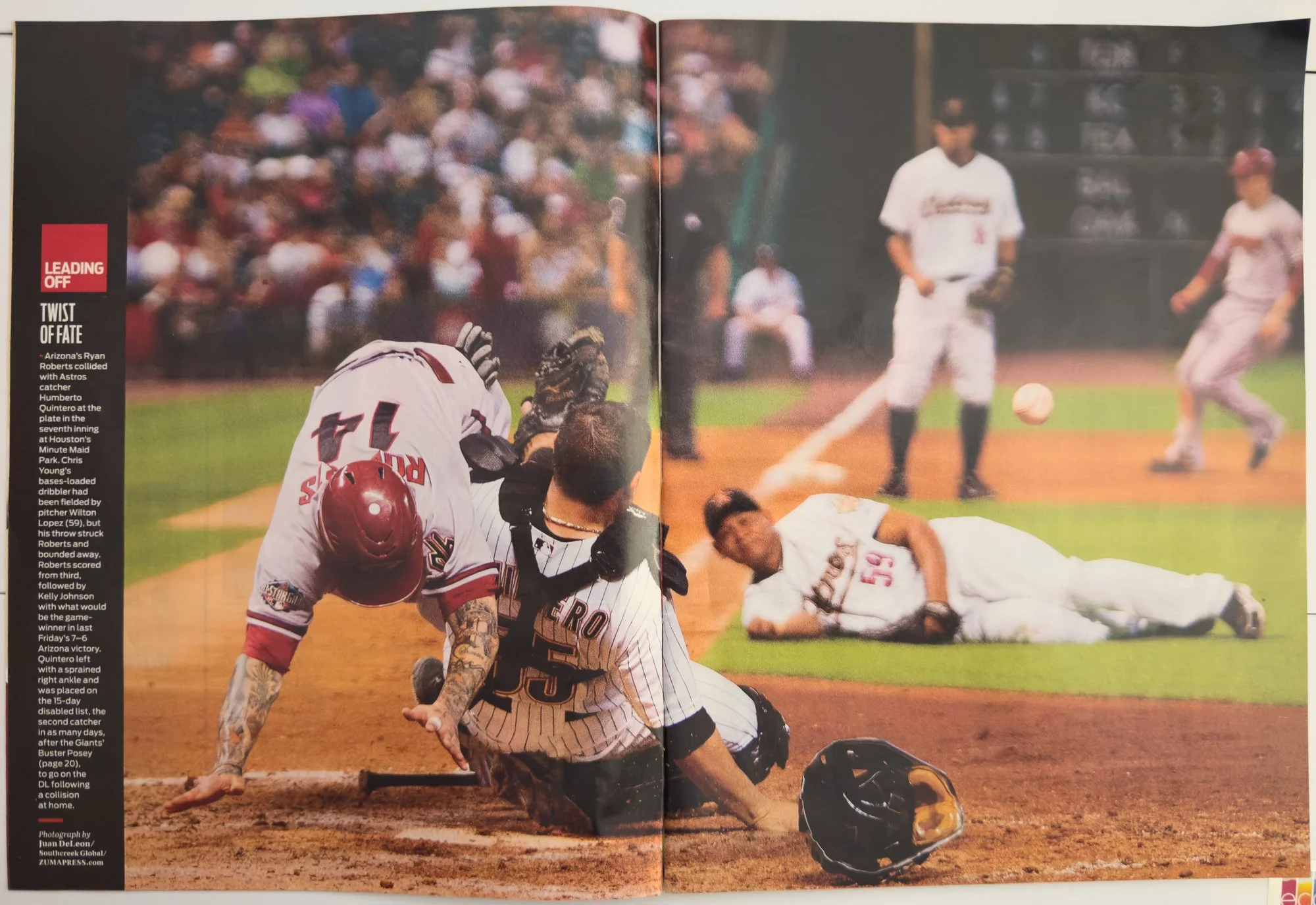 A baseball game in progress showing a player sliding into home plate, a catcher from the opposing team blocking the plate, and an umpire watching the play. There are players and spectators in the background.