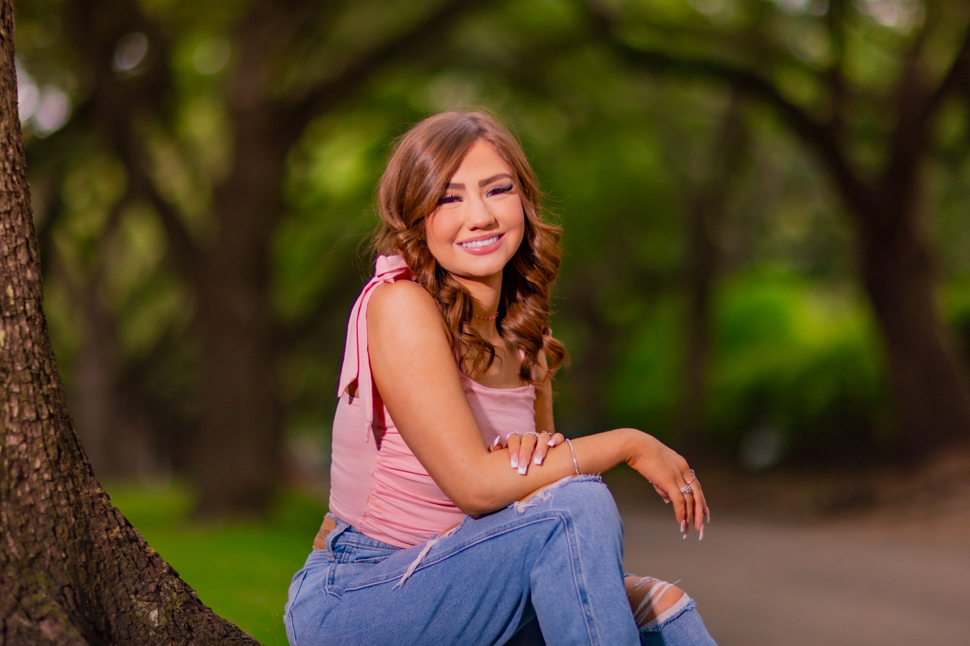 Young woman with curly brown hair sitting on a tree branch in a park, smiling with closed eyes, wearing a pink top and ripped jeans.