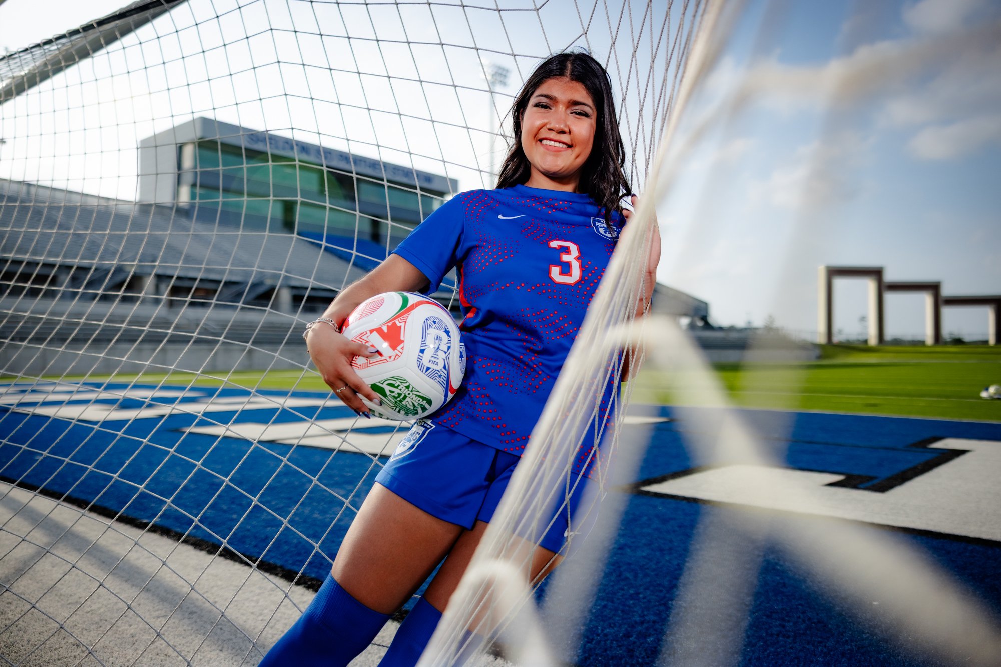 A young woman in a blue soccer uniform standing inside a soccer goal net, holding a soccer ball, at a sports stadium during daytime.