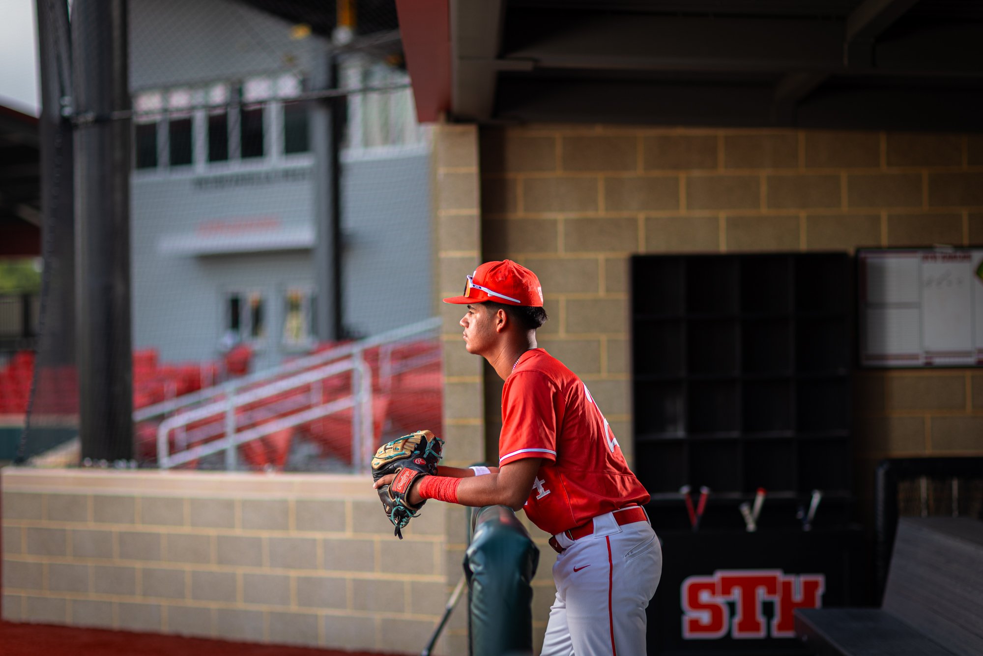 A young baseball player in a red uniform and cap standing on a dugout bench, holding a baseball glove, looking to the side. The background shows part of the baseball field and dugout.