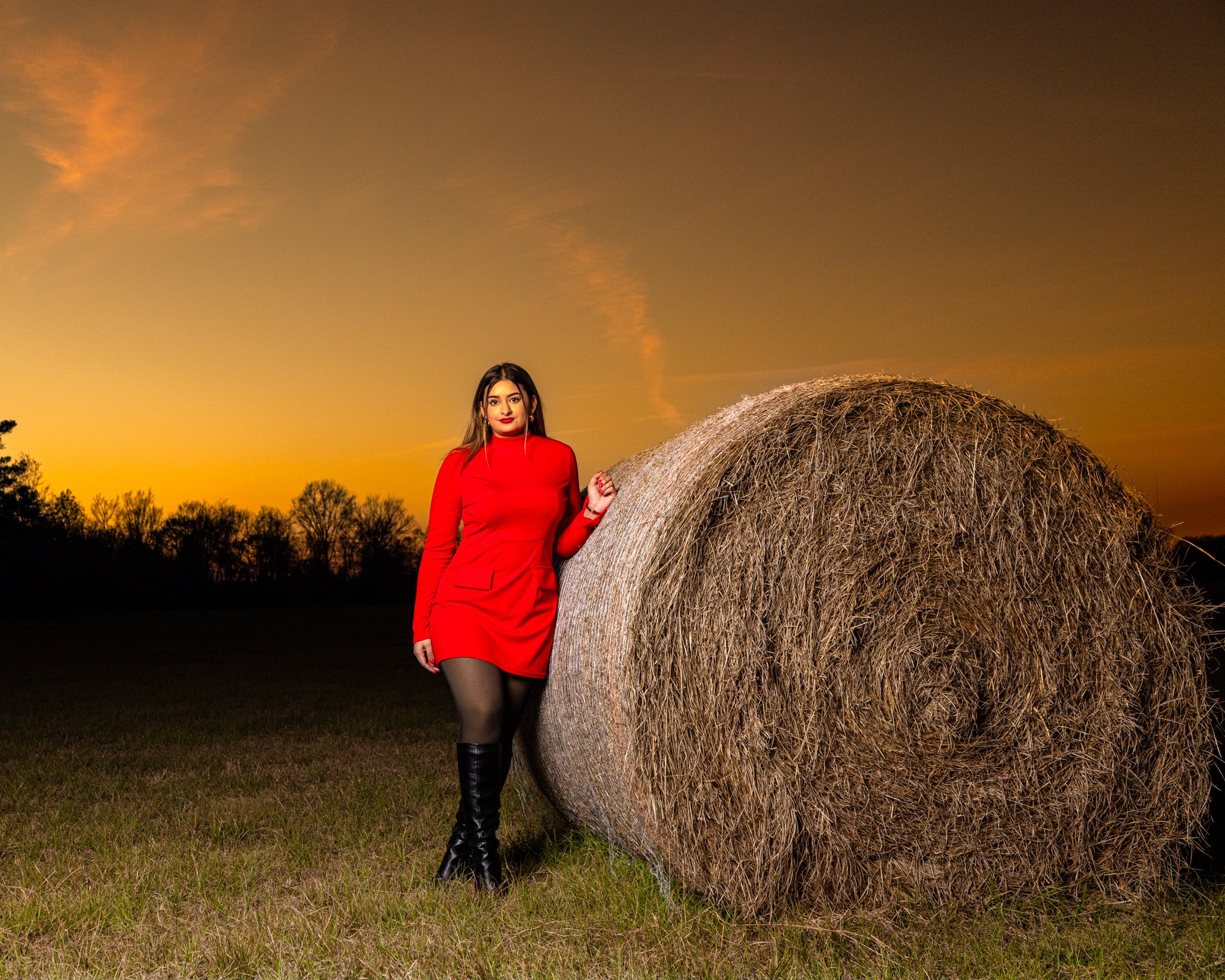 A woman dressed in a red outfit standing next to a large hay bale during sunset in a rural field with a tree line in the background.