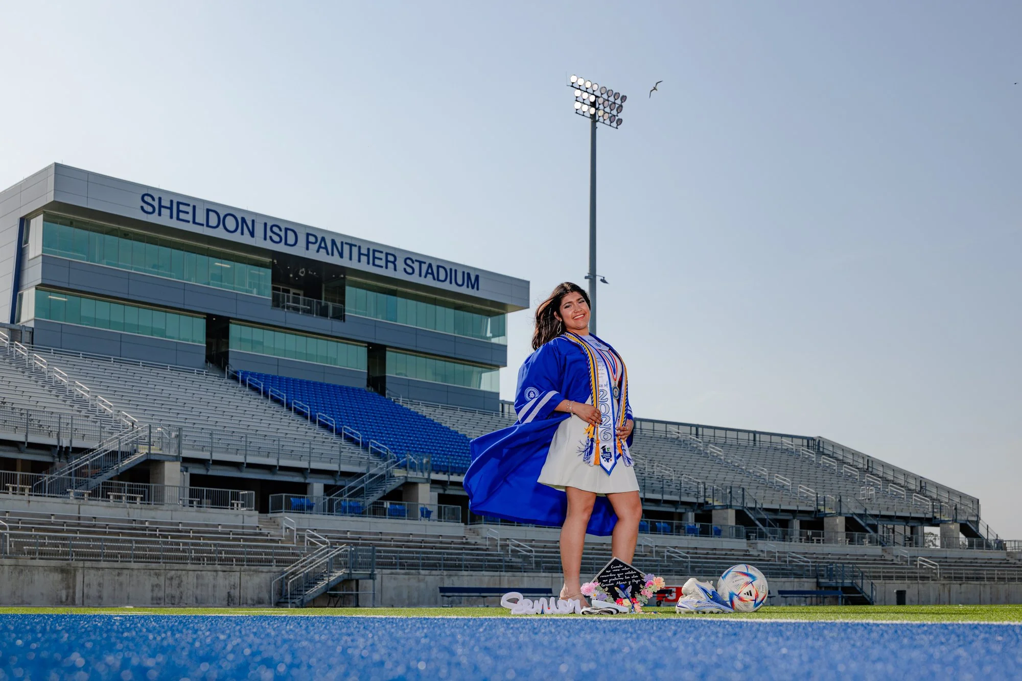 A young woman dressed in a graduation gown and sash standing on a football field at Sheldon ISD Panther Stadium, with flowers, shoes, and soccer balls on the ground at her feet, during daylight with clear sky.