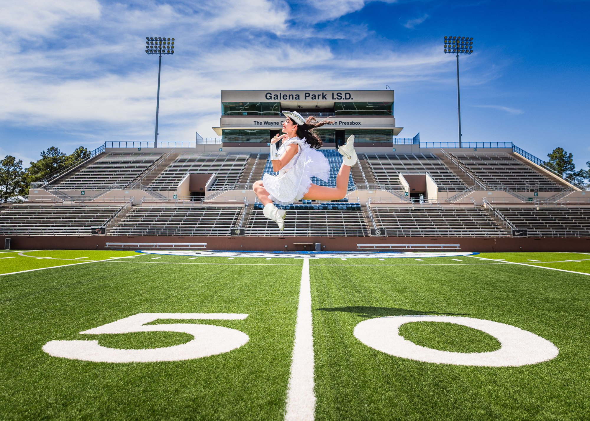 A young woman in white dress and hat jumping in the center of a football field with empty bleachers and stadium lights in the background.