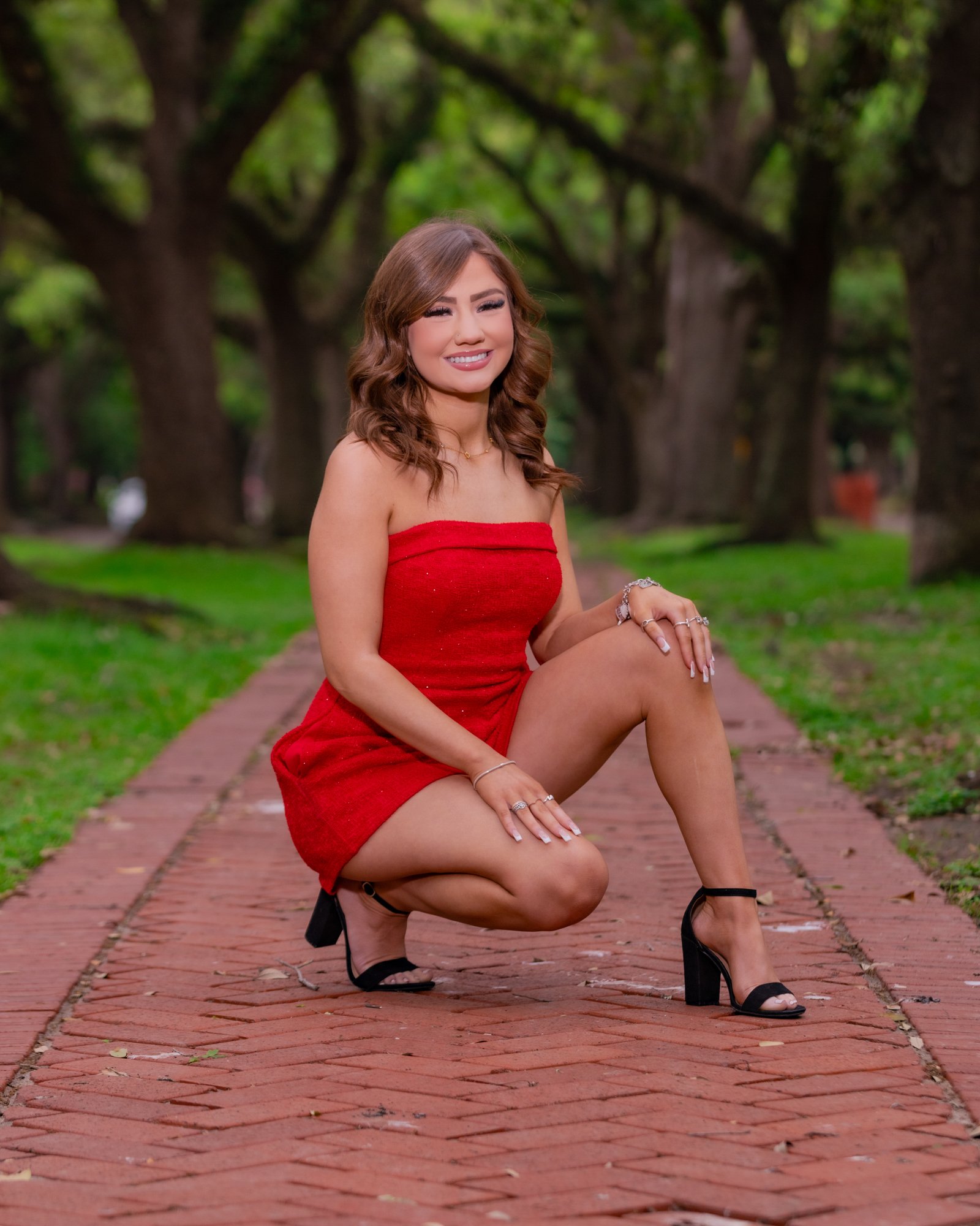 A woman in a red dress squatting on a brick pathway in a park with trees in the background, smiling and winking at the camera.