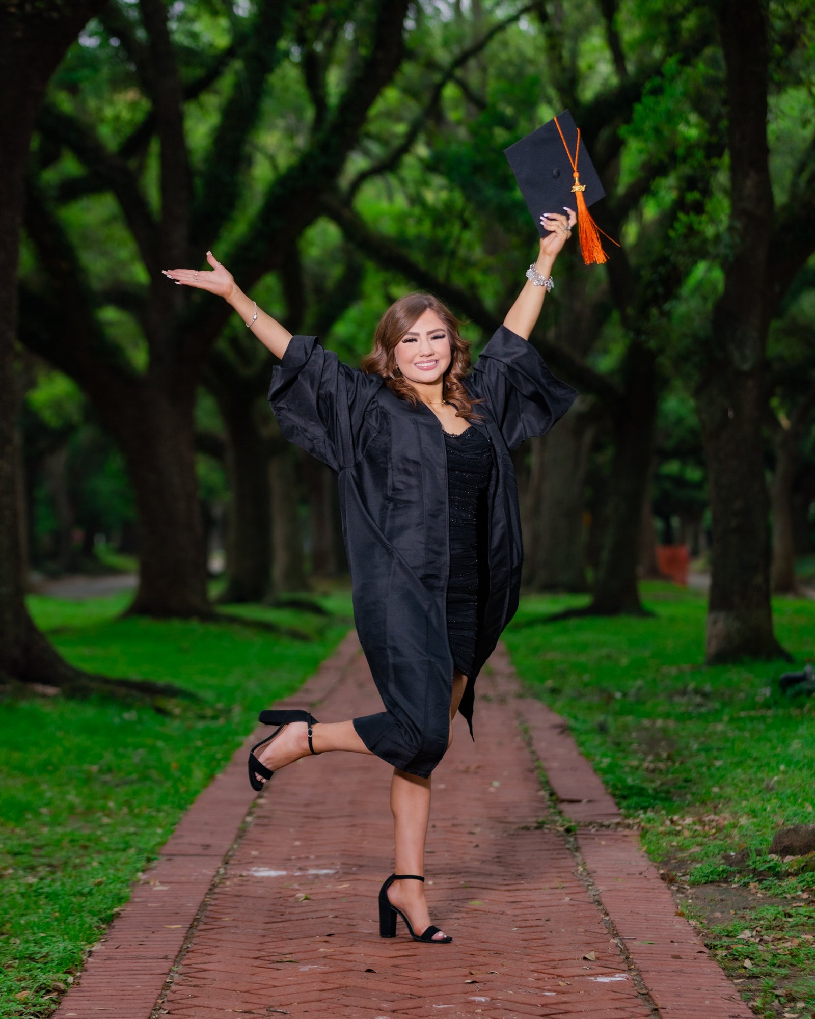 A woman in a graduation gown is celebrating outdoors in a park. She is holding a black graduation cap with an orange tassel and a diploma, smiling, with one leg bent and arms raised. The background features green trees and a brick pathway.