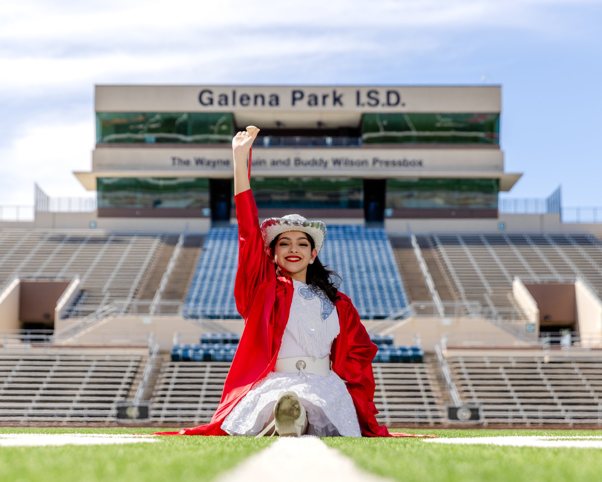 A young girl with a wide smile, dressed in a white dress and red jacket, is sitting on the grass in a stadium, holding a small trophy above her head. The stadiums seating and a sign that reads 'Galena Park I.S.D.' are visible in the background.