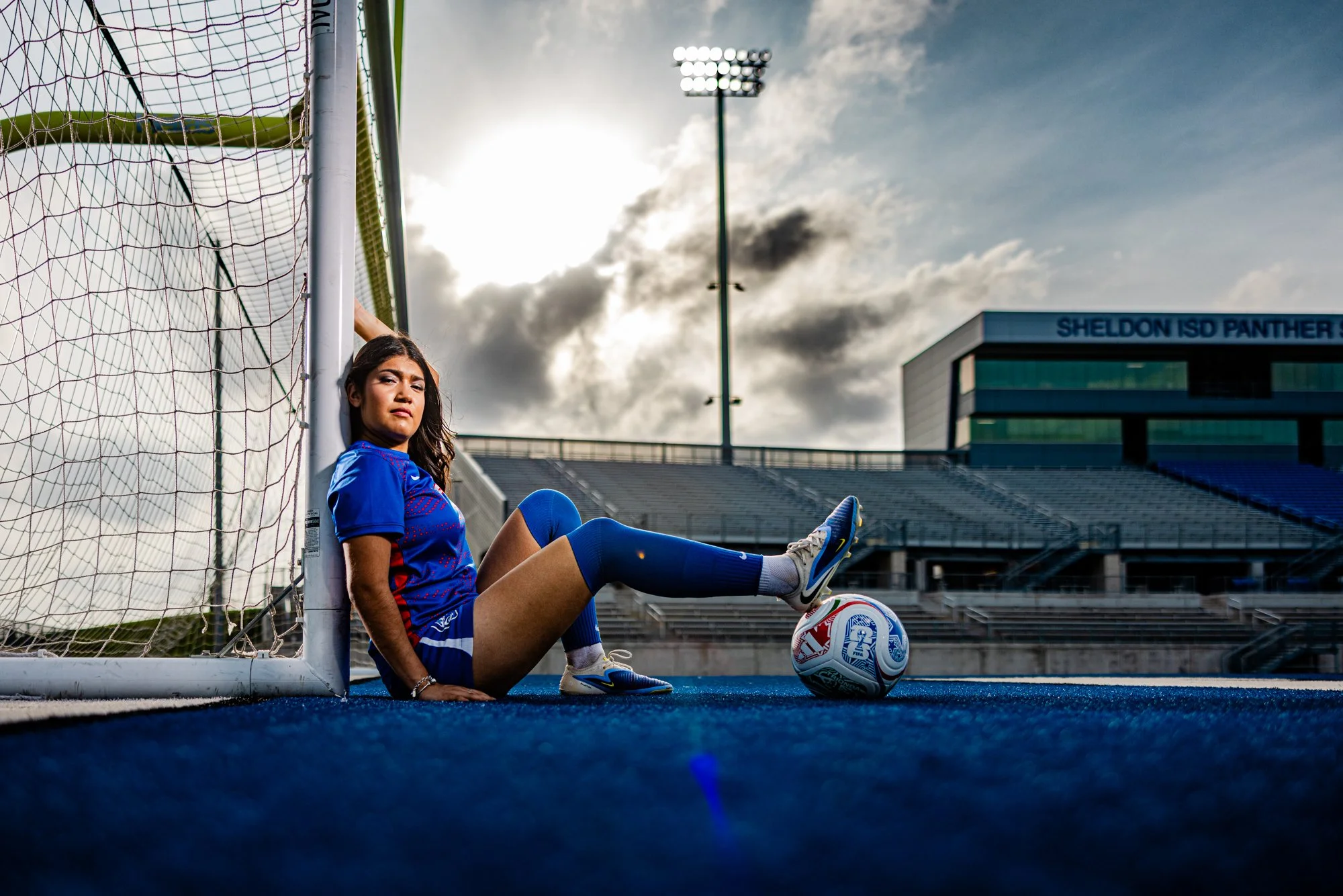 A female soccer player sitting on the blue artificial turf field next to the goalpost, with her back against it. She is wearing a blue jersey and shorts, and has one leg extended and resting on the soccer ball, which is positioned near her foot. The 