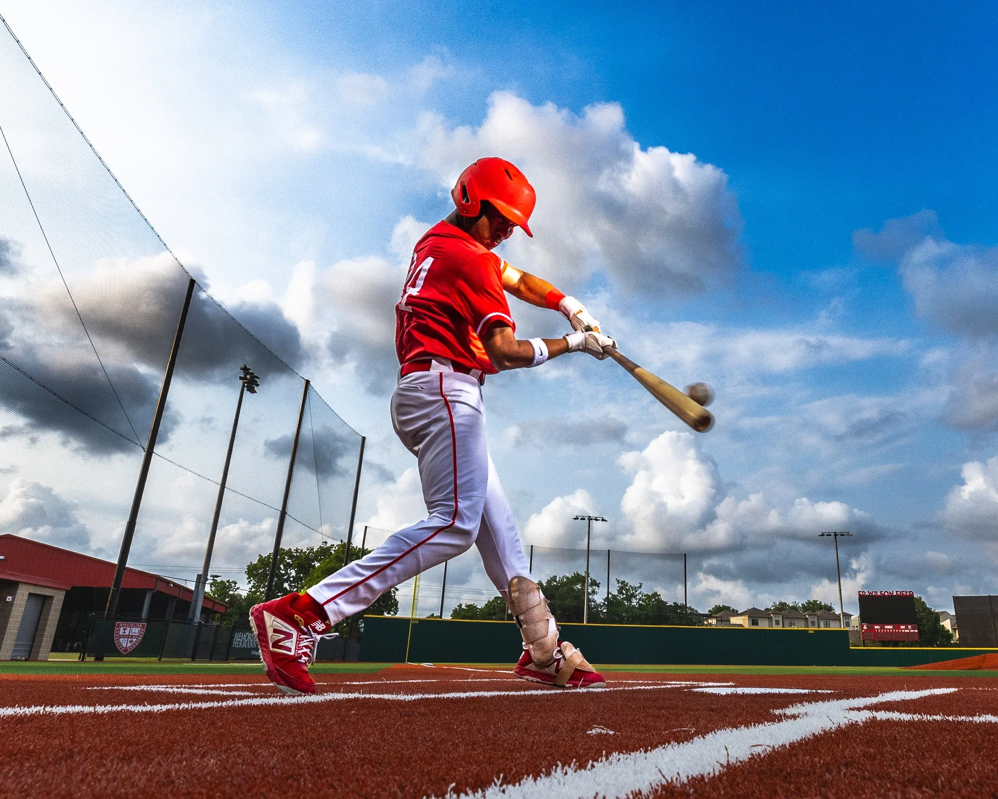 A baseball player wearing a red helmet, red shirt, and white pants is running on a baseball field. The player holds a bat and wears a tan leg guard. The sky is partly cloudy, and there are tall fences, stadium lights, and buildings in the background.