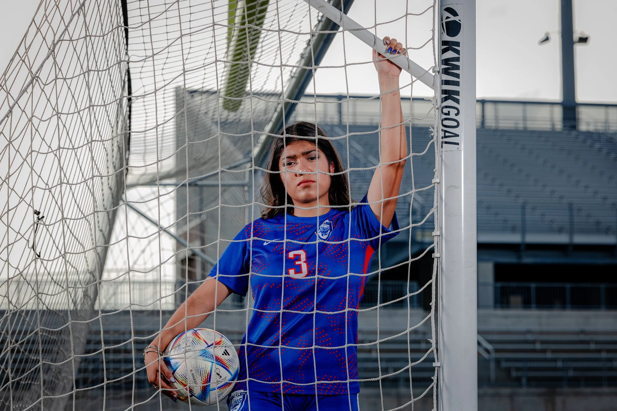 A female soccer player dressed in a blue jersey with the number 3, standing behind a goal net and holding a soccer ball, on a soccer field outdoors.