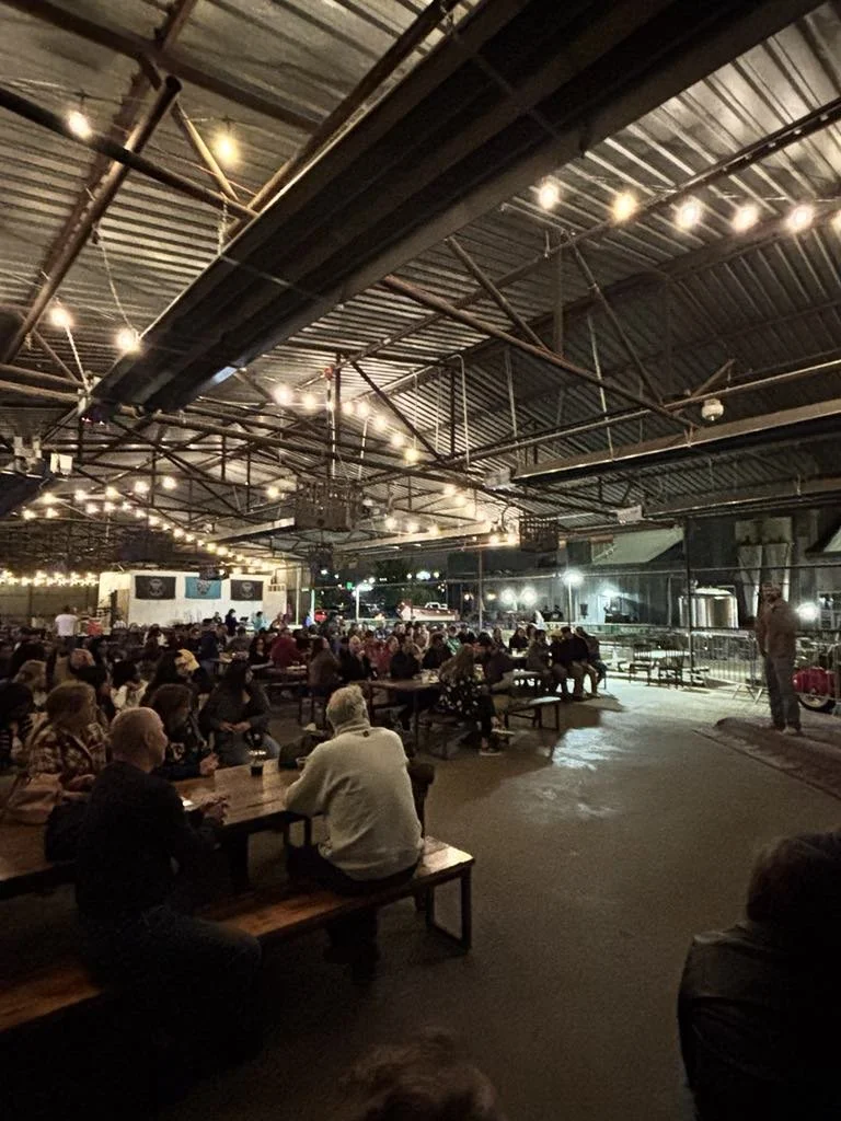 People sitting at picnic tables in a covered outdoor area at night, with string lights overhead and a stage or performance area in the background.