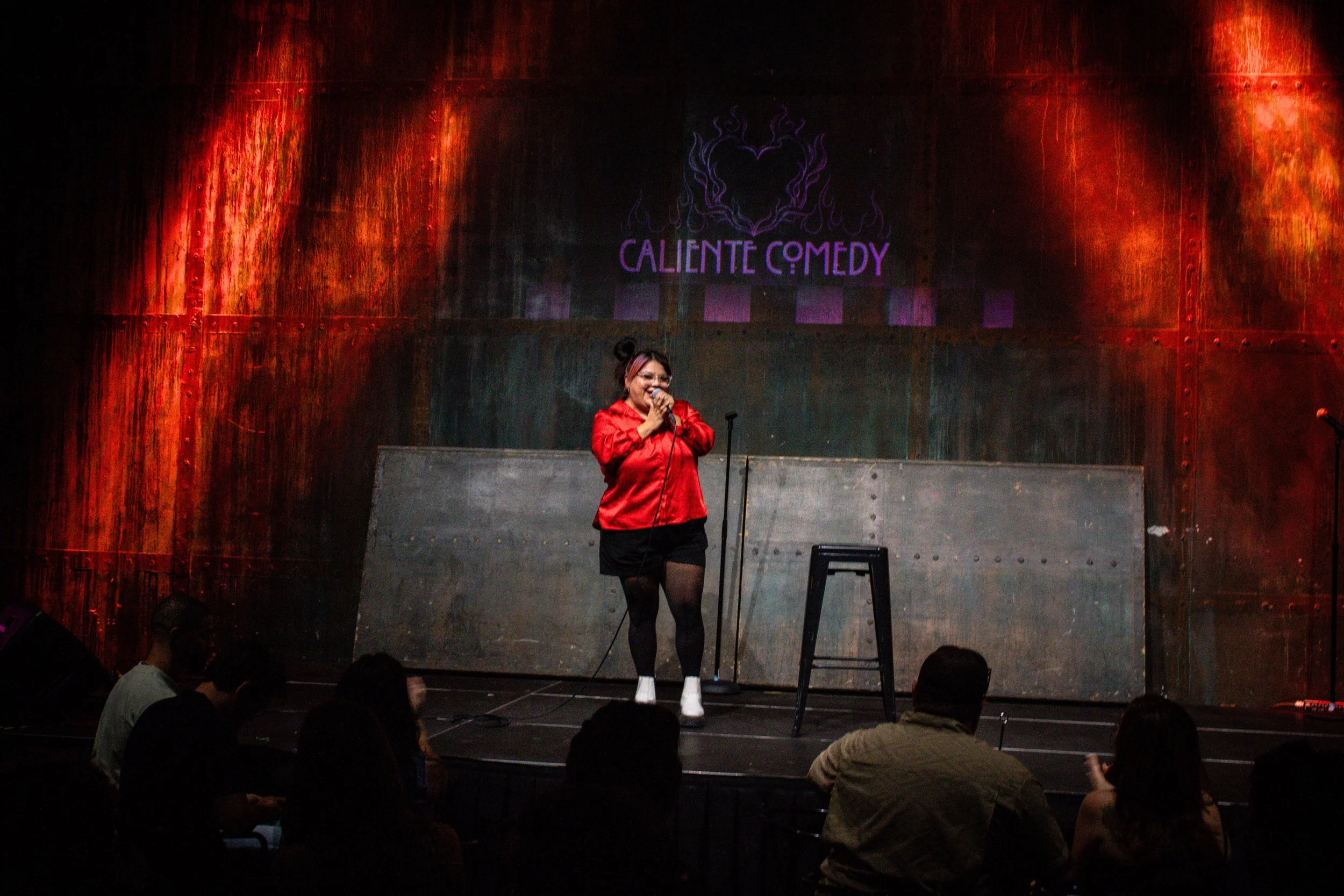 A comedian performing on stage at Caliente Comedy, with a red jacket, holding a microphone, in front of a dark background with a purple sign that says 'Caliente Comedy' and a stylized flame logo.