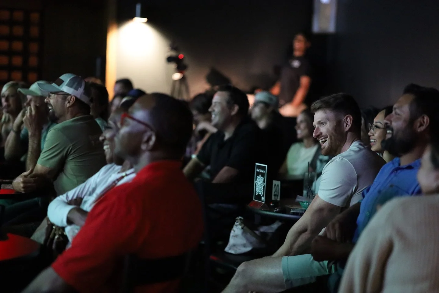 Audience members seated and laughing during a comedy or entertainment show in a dark venue.