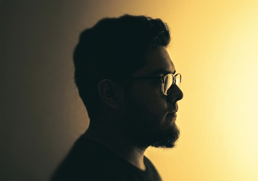 Silhouette of a man with glasses looking to the right, illuminated by warm light in a side profile view.