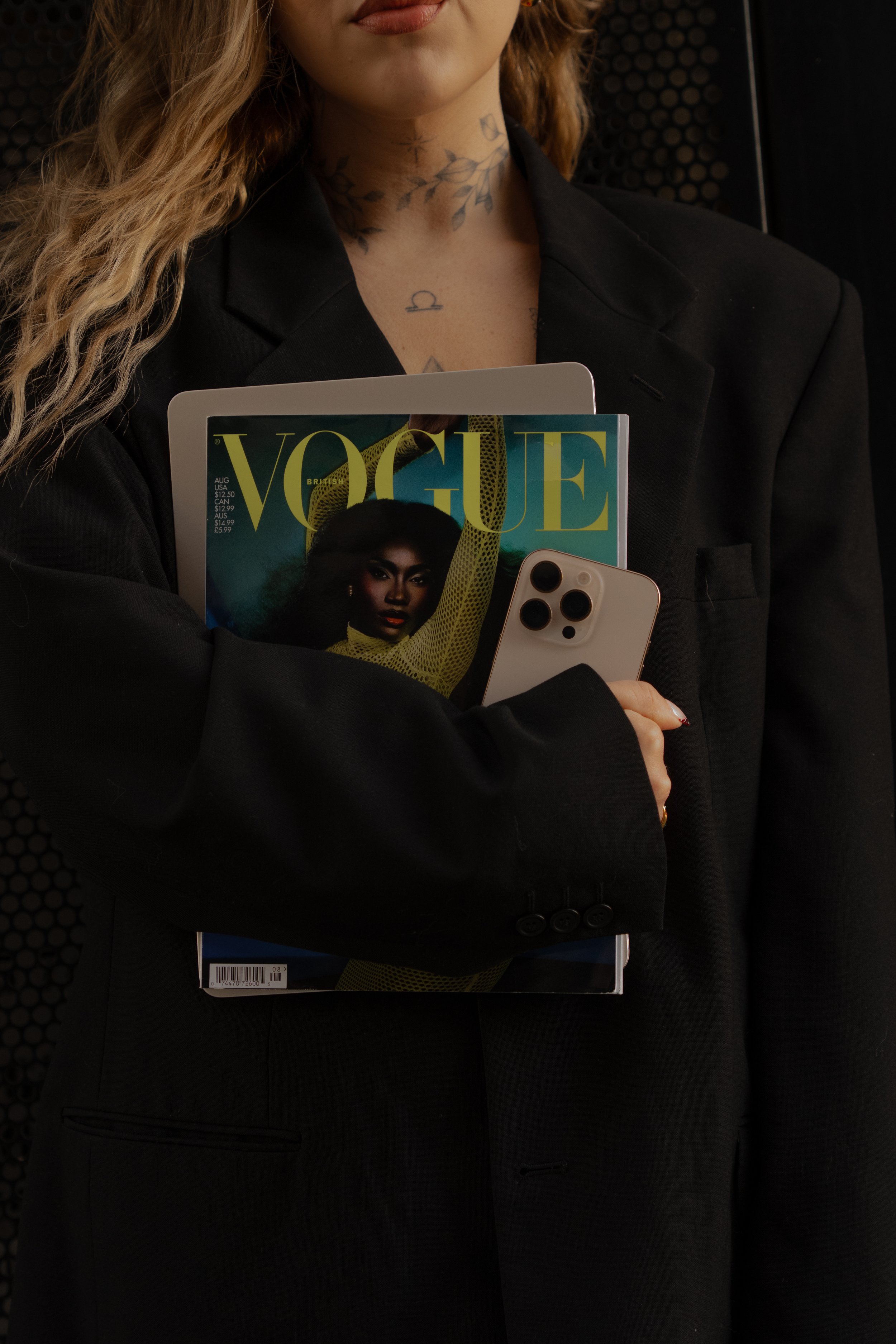Close-up of a woman in a black blazer holding a Vogue magazine, a smartphone, and a notebook, with visible tattoos on her neck and chest.
