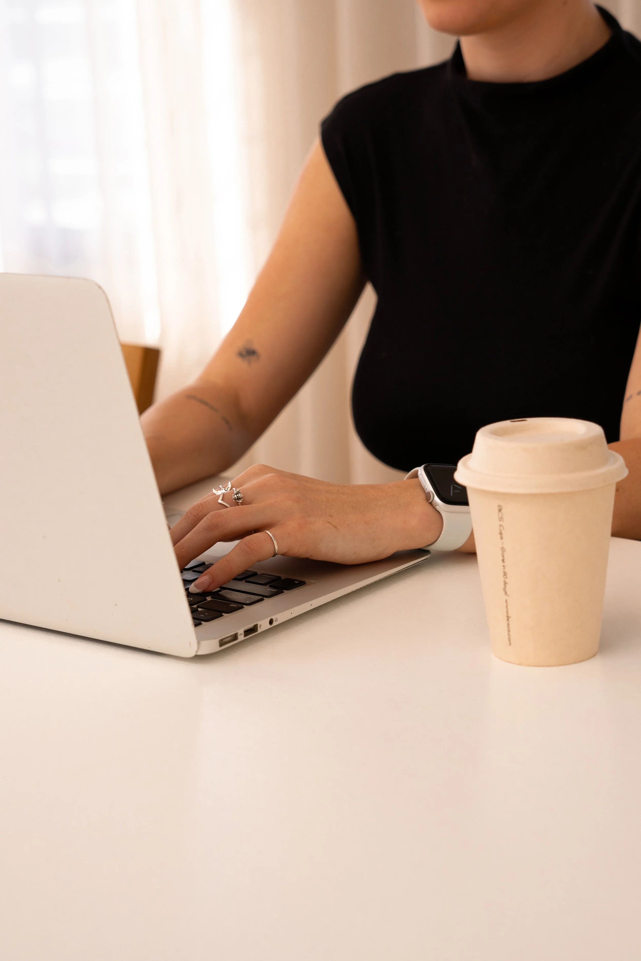 A person working on a laptop, wearing a black shirt, with a disposable coffee cup on a white table.
