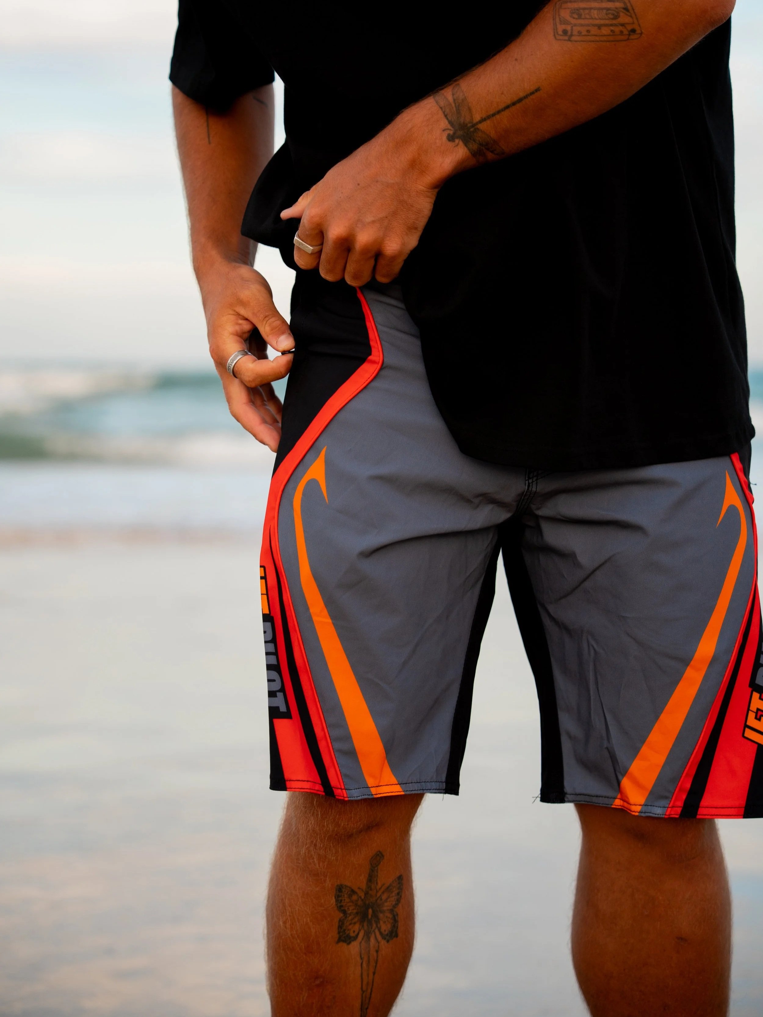 Person wearing black and gray shorts with red and orange accents, standing on a beach with ocean waves in the background, showing tattoos on their arm and leg.