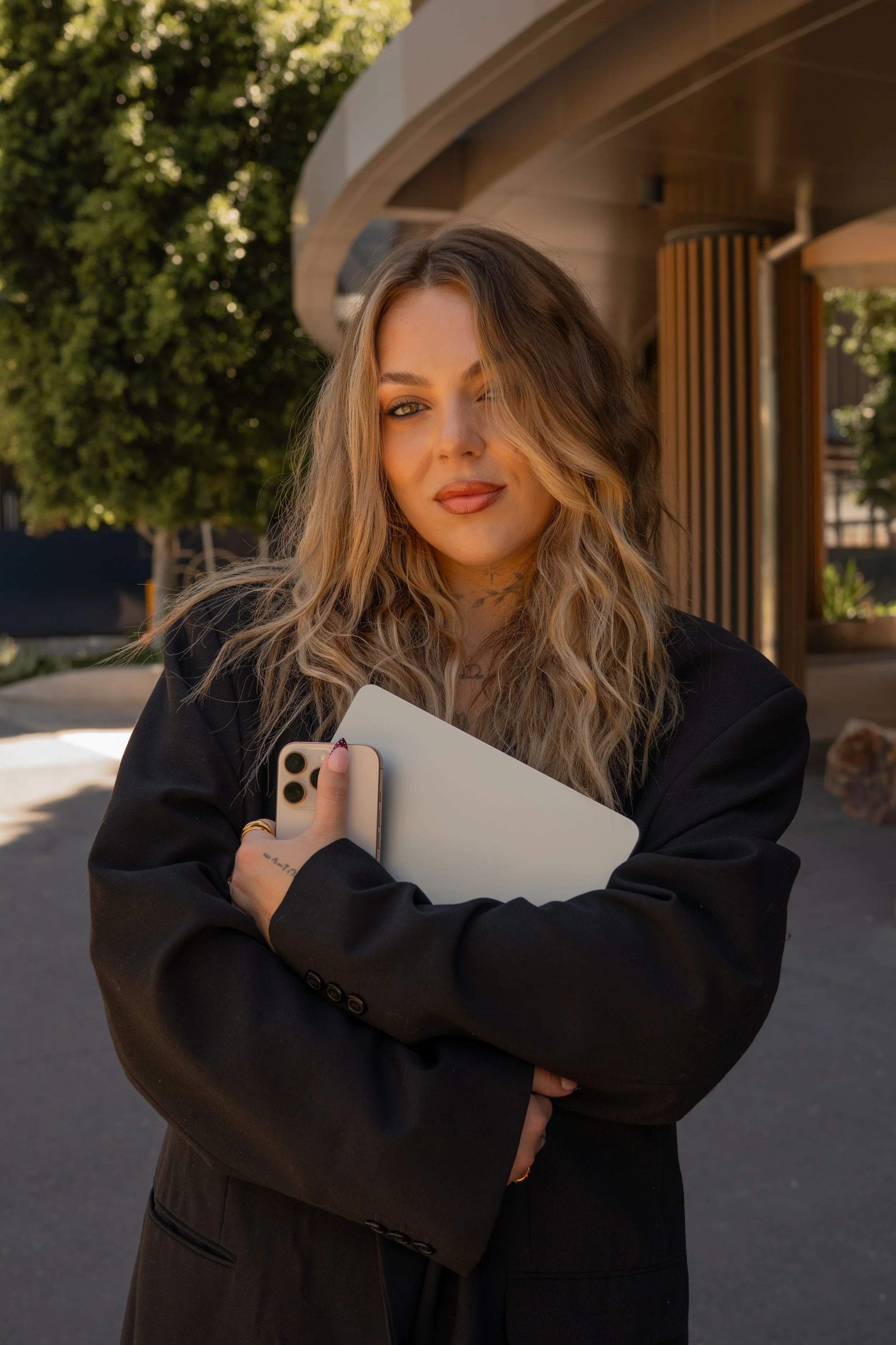 A woman with long wavy blonde hair, winking and smiling, holding a smartphone and a white book or notebook, standing outdoors with trees and a modern building in the background.