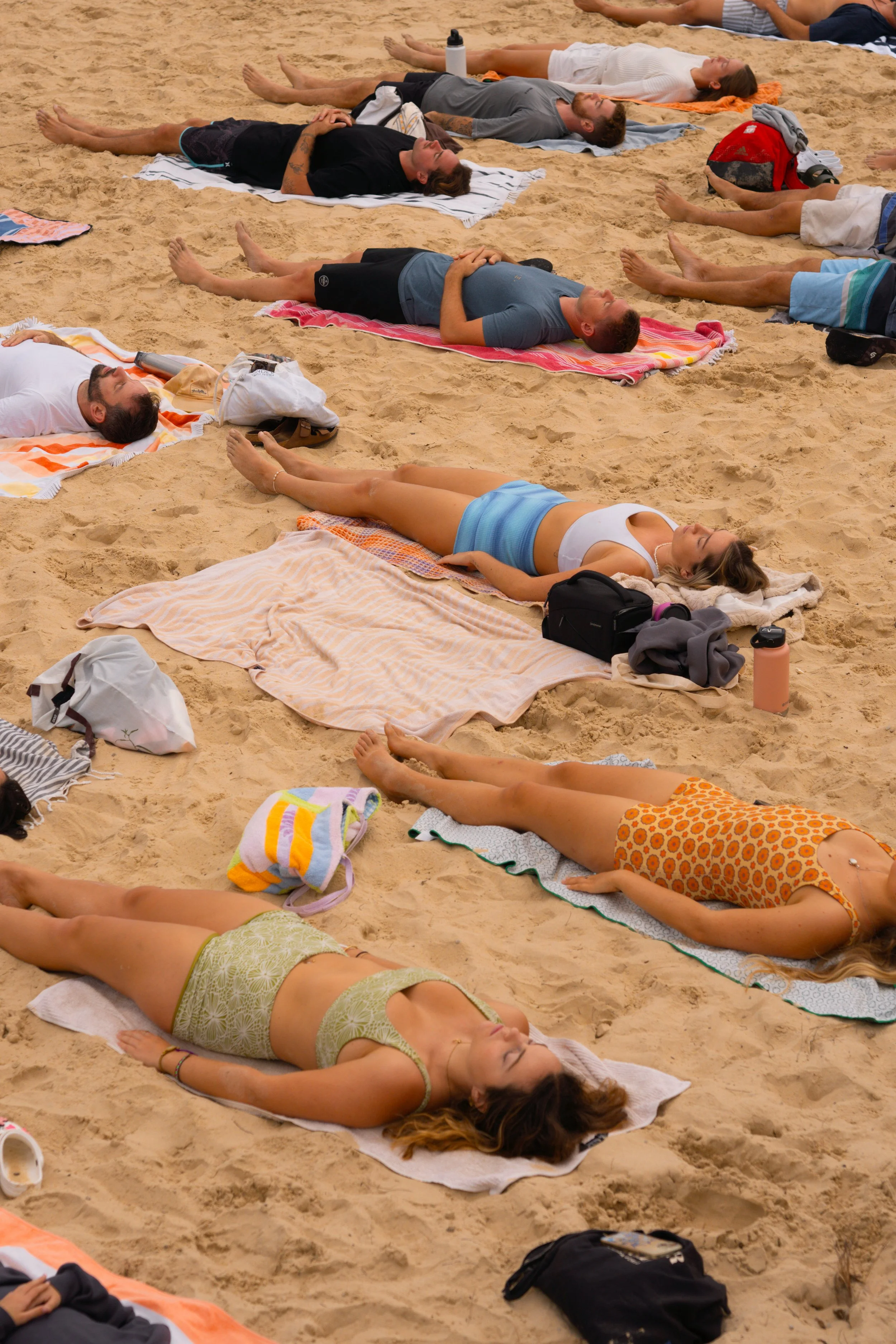 Multiple people lying on sand at the beach, some on towels, taking a nap or relaxing, with their eyes closed.