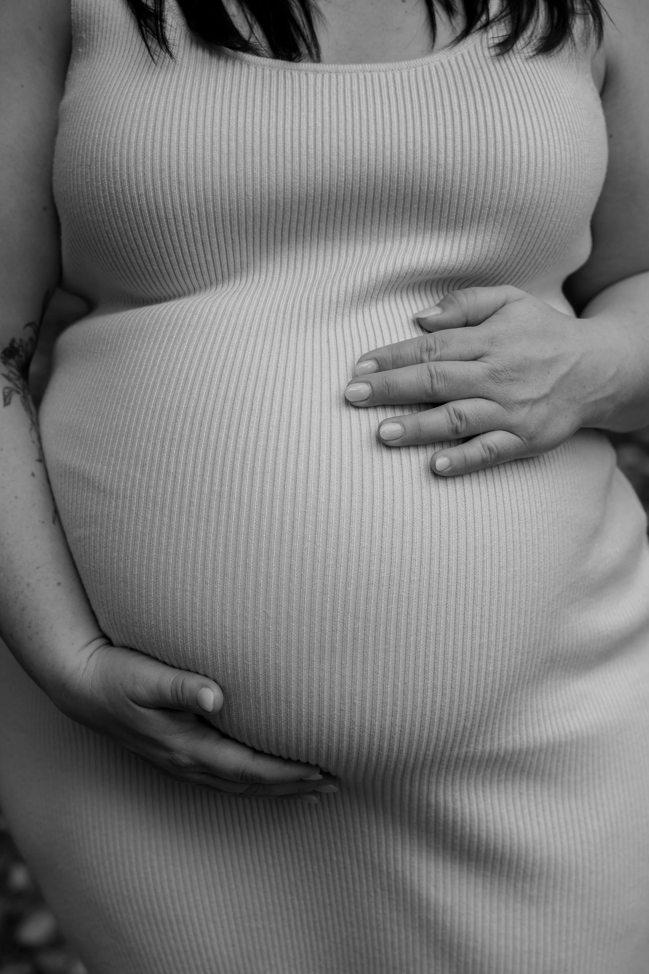 Close-up black and white photo of a pregnant woman wearing a striped sleeveless top, cradling her belly with her left hand.