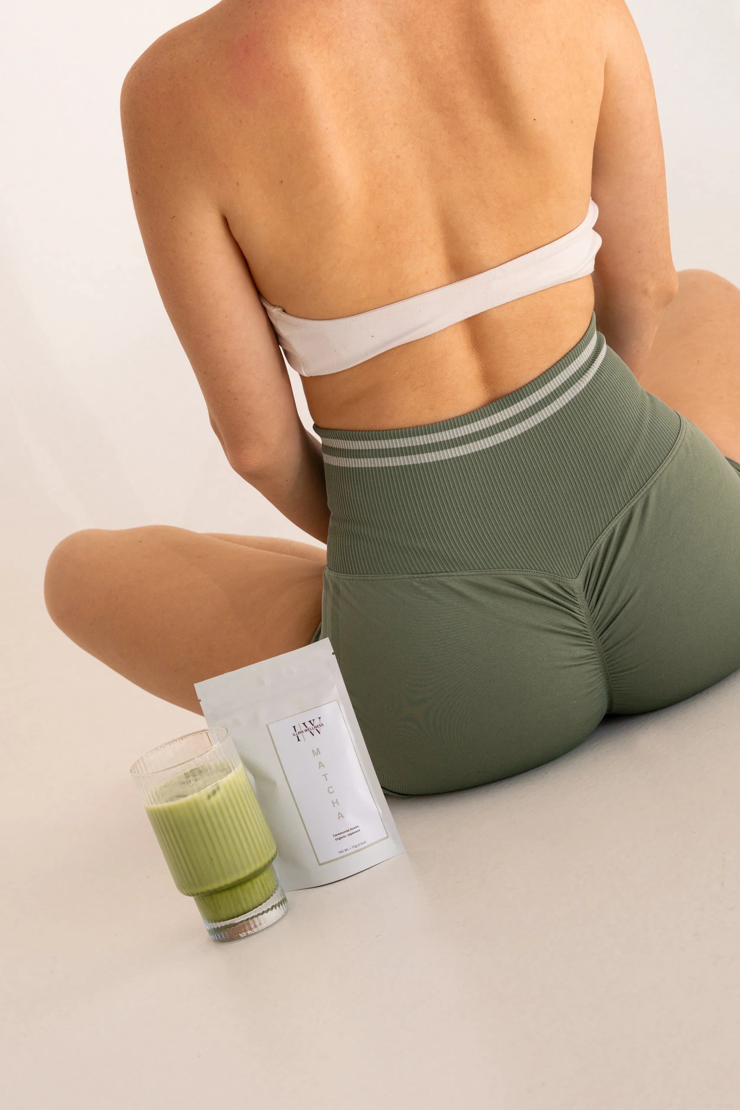 A woman wearing green workout leggings and a white strapless top, sitting on the floor with her back to the camera. In front of her are a glass of green beverage and a white tea packet labeled 'Matcha'.