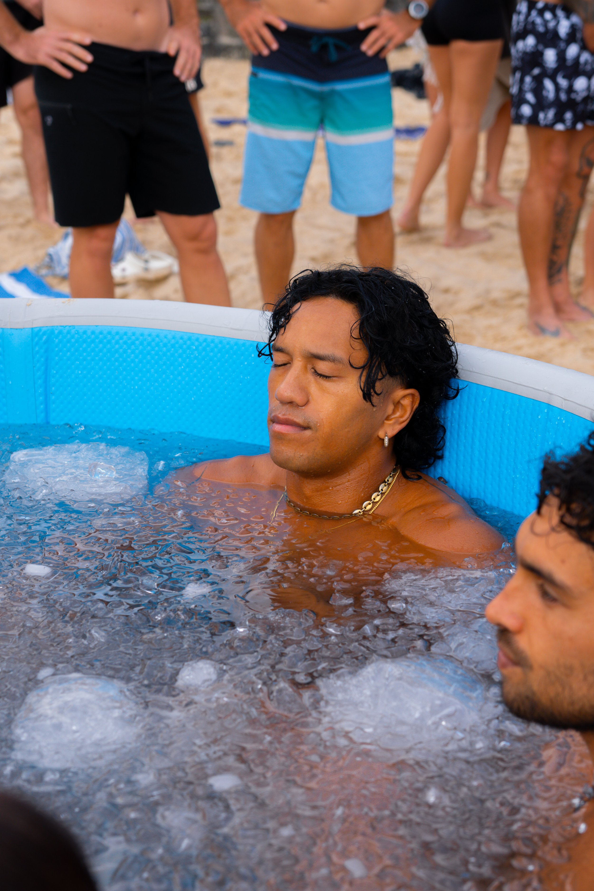 People standing on a sandy beach, wearing swimwear, with a man relaxing in a small inflatable pool.