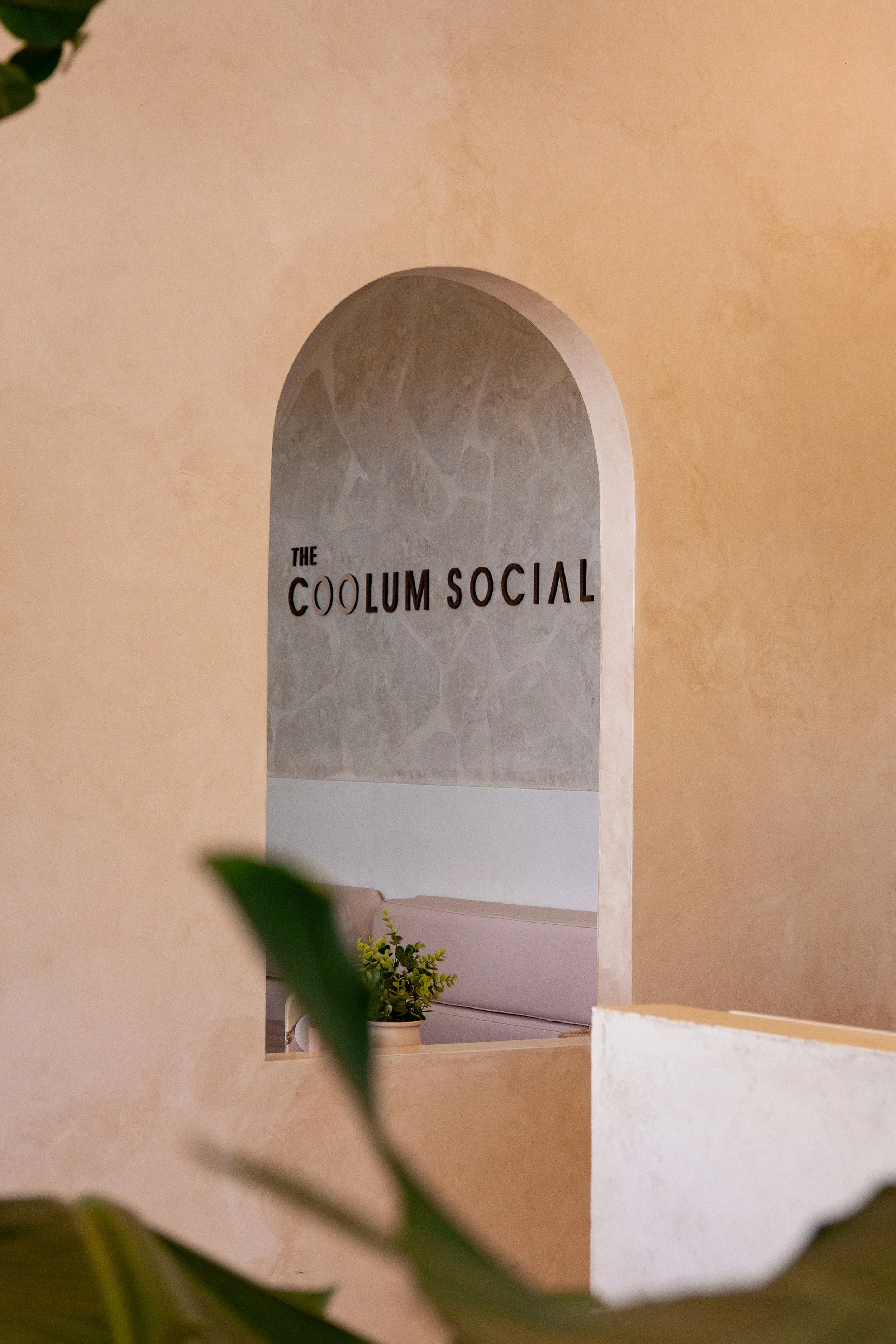 Interior of a stylish space with a decorative arched wall featuring the words 'The Coolum Social' and some greenery in the foreground.