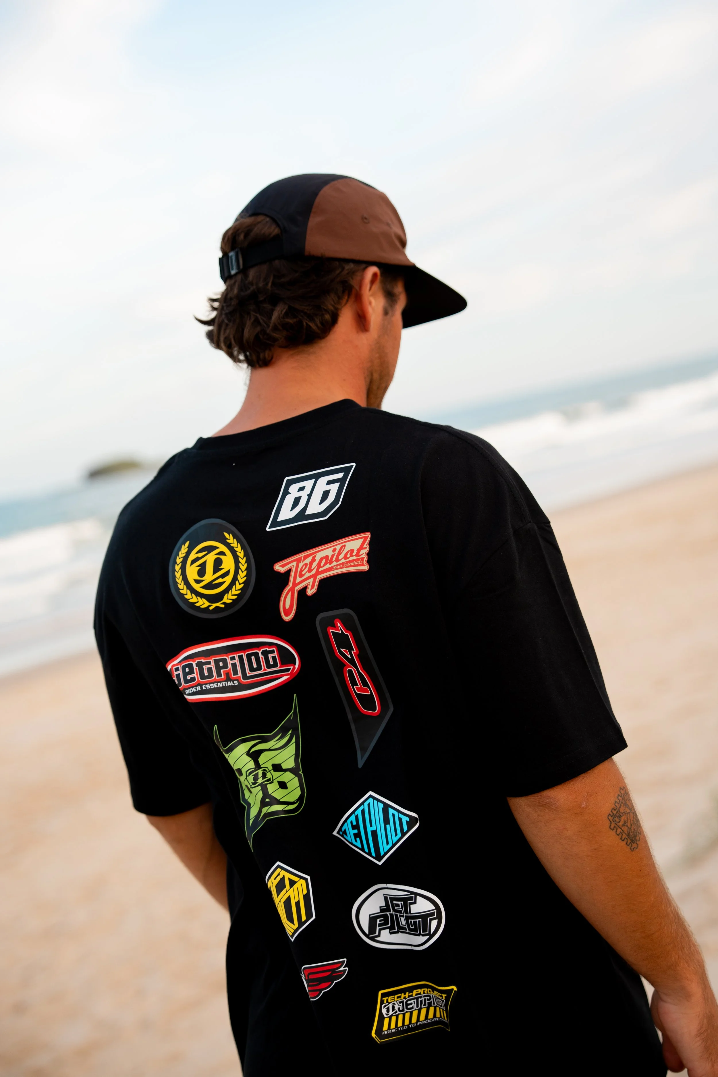 Person standing on a beach with their back to the camera, wearing a black t-shirt with multiple racing stickers and a brown and black cap.